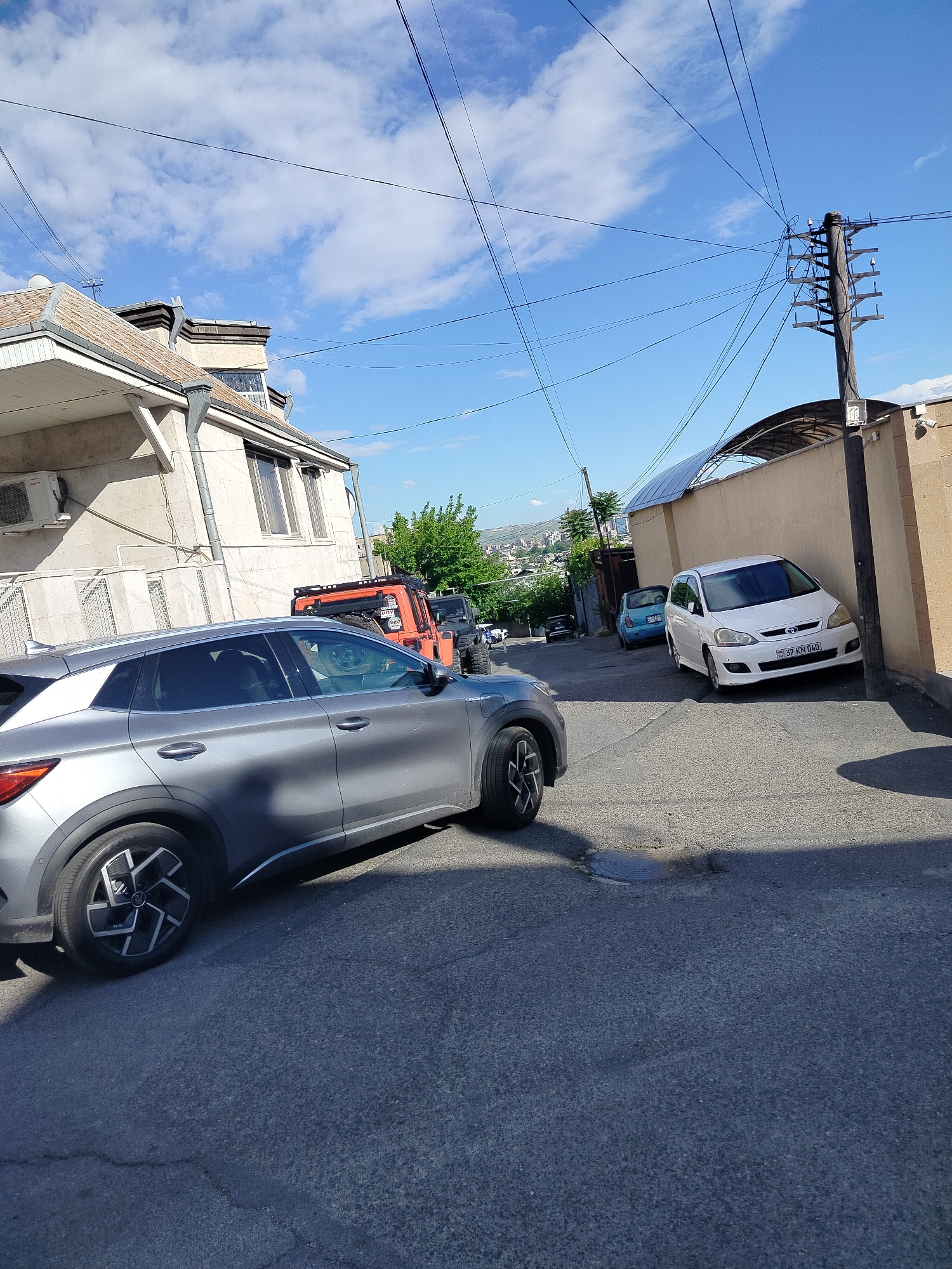 The image depicts a street scene in Yerevan, Armenia. The street is narrow, paved with asphalt, and has several parked cars. In the foreground is a gray SUV, partially obscuring an orange Jeep further down the street. A white Toyota is parked against a tan-colored wall with a dark metal awning. A light blue Fiat is parked behind it. An electrical pole is visible near the Toyota. Buildings line both sides of the street. A distant view of hills and city buildings is visible between the buildings. The sky is blue with scattered white clouds.