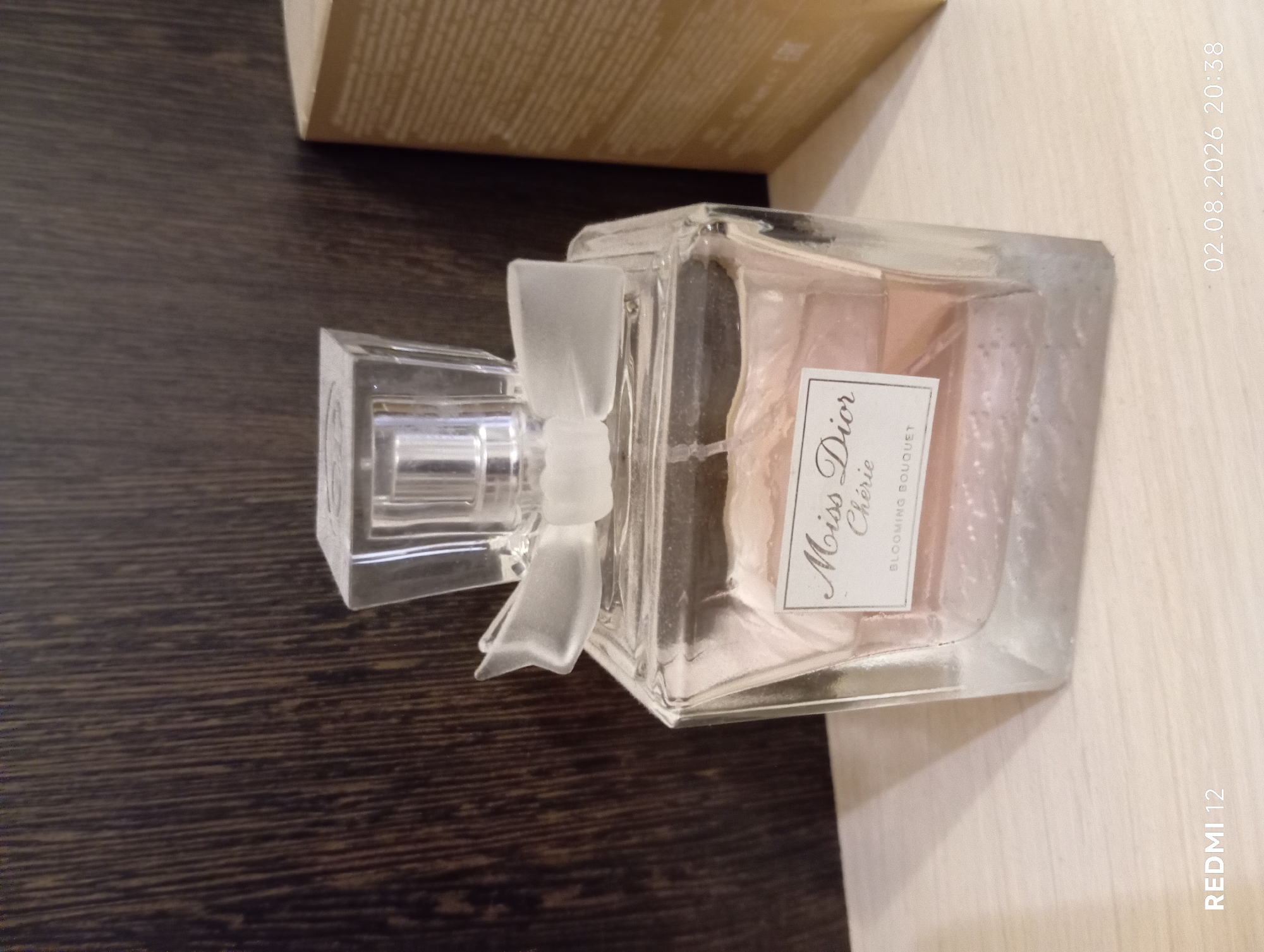 A close-up, eye-level shot features a glass perfume bottle with a pink liquid and a bow-shaped atomizer. The bottle is labeled "Miss Dior Chérie Blooming Bouquet." It sits on a light-colored wooden surface. In the background, a dark wood-paneled wall is visible, along with a corner of a tan cardboard box. The lower right corner displays a timestamp: "02.08.2026 20:38" and "REDMI 12" is in the lower left corner. The image appears to be taken indoors, possibly in an apartment or room. There are no people or animals present. The scene is static and appears to be a product photograph or personal item display. The location is not specified in the image itself, but the presence of the timestamp and phone model information may suggest it was taken by a user in Russia, potentially in Oryol.