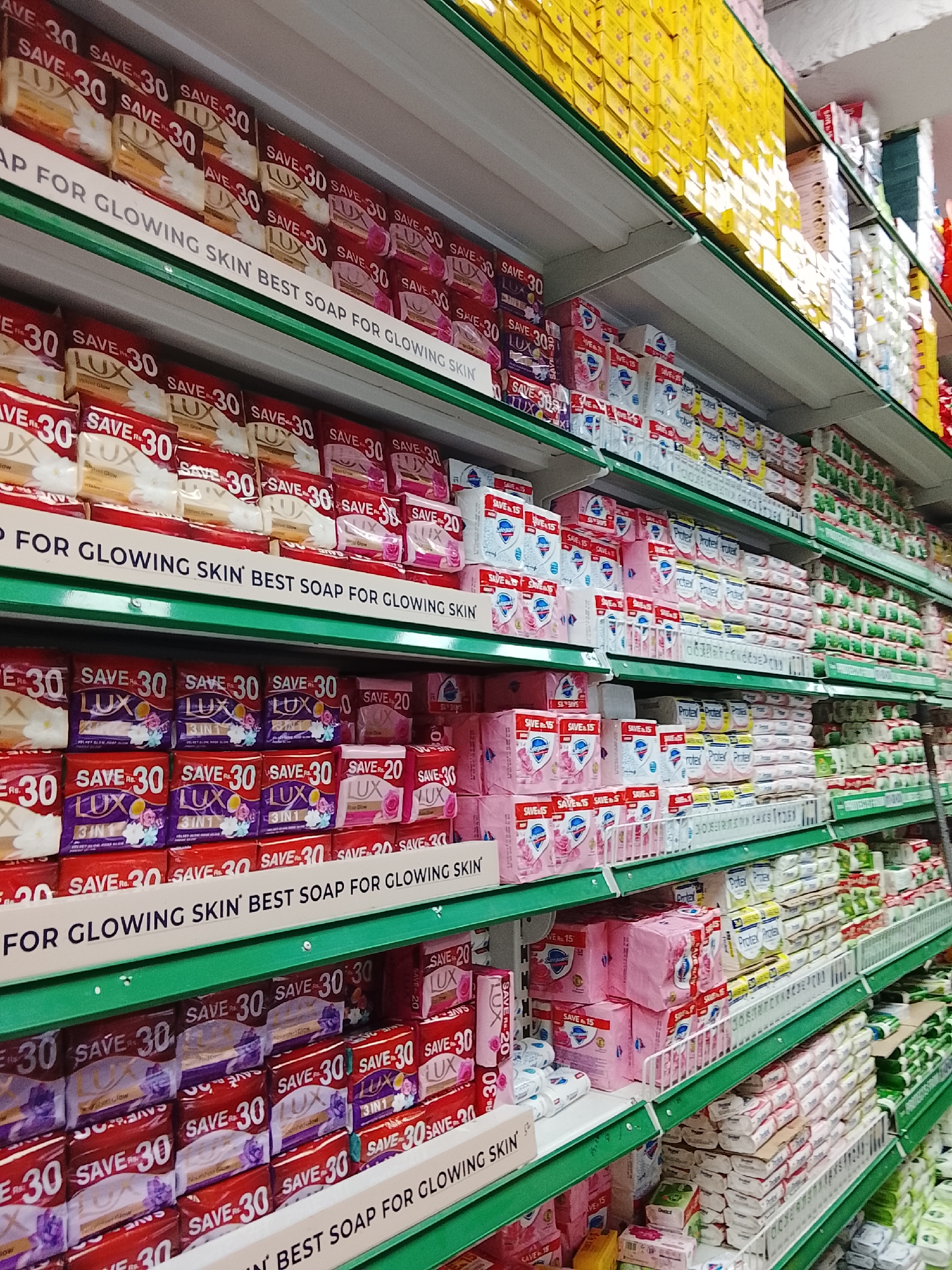 The image depicts shelves in a retail environment, likely a store or supermarket. The shelves are stocked with various packaged goods, notably soap bars labeled "LUX" with promotions such as "SAVE 30," suggesting a discount or deal. There are also sanitary pads with "SAVE 15" promotions. The signage indicates the soap is "BEST SOAP FOR GLOWING SKIN." The shelves are green and organized with rows of products, and they are located in Gulgasht Colony, Pakistan. The image contains no people.
