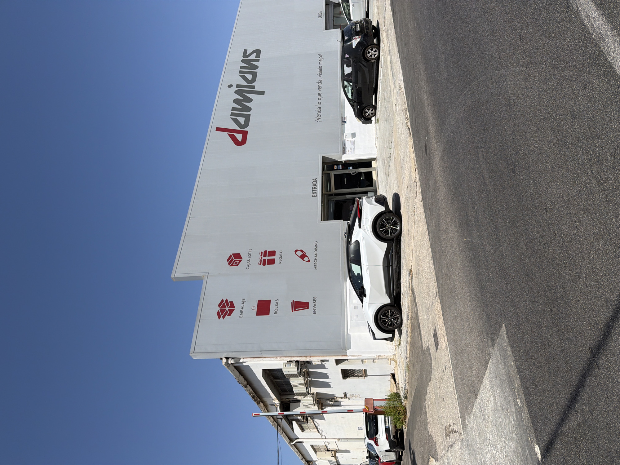 In the Polígon Industrial De Son Castello, Spain, a white building with a large red logo that reads "damians" is visible under a clear blue sky. The building's facade is adorned with various red icons representing packaging, gift boxes, bags, cups, and a pen, with accompanying Spanish text for "EMBALAJE" (packaging), "CAJAS LOTES" (batch boxes), "BOLSAS" (bags), "REGALO" (gift), "ENVASES" (containers), and "MERCHANDISING." To the left of the building, an older, white structure with windows and air conditioning units is attached. A white barrier gate stands to the far left. In front of the building, a white SUV is parked. Adjacent to the white SUV is a black hatchback car. A modern white SUV is parked in front of the building's entrance, which is marked "ENTRADA" (entrance). The slogan "¡Venda lo que venda, vistalo mejor!" (Sell whatever you sell, dress it better!) is written on the building's exterior. A sign displaying "HORARIO" (schedule) with opening and closing times is visible near the entrance. The ground in front of the building is paved. The image captures a commercial or industrial area on a sunny day.
