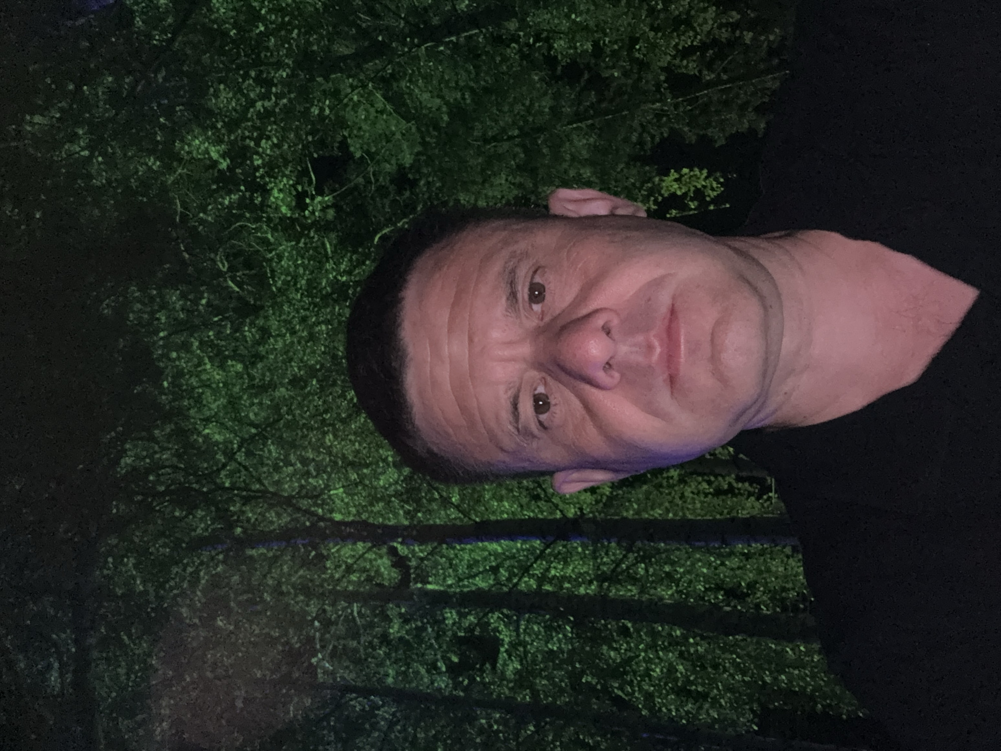 A selfie of a middle-aged white man is taken outdoors at night in front of lush green trees. The man is wearing a black shirt and looking directly at the camera. The lighting is dim, with a green hue illuminating the trees and a subtle blue and pink cast on the man's face. The background consists of dense foliage and tree trunks, suggesting a natural or park-like setting. The man's facial features are prominent, with visible wrinkles and a serious expression. The overall atmosphere is somewhat dramatic due to the lighting and background.