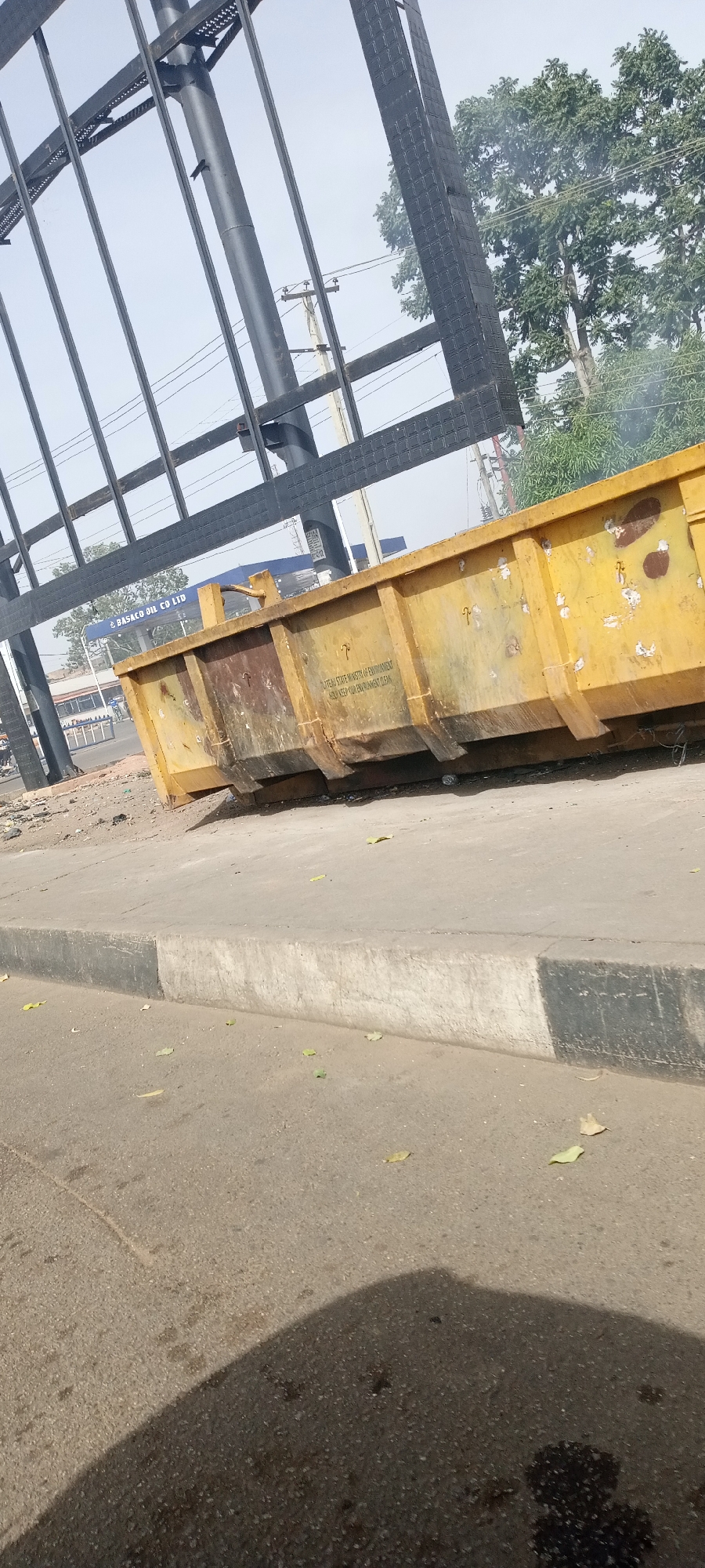 The image captures a static roadside scene in Jos, Nigeria, during bright daylight hours under a clear sky. The primary subjects are a large, weathered yellow industrial dumpster or skip bin and a prominent black metal billboard frame.
The dumpster is positioned on a dirt verge alongside a concrete curb and an asphalt road. It shows signs of use with dirt, rust, and wear. Visible text on its side reads, "PLATEAU STATE MINISTRY OF ENVIRONMENT" and "HELP KEEP OUR ENVIRONMENT CLEAN," indicating its purpose for waste management and environmental awareness.
Above and behind the dumpster, a substantial black metal framework of a billboard stands tall. One of the supporting poles of this structure has a small, dark bird perched on it. In the background, through the billboard frame, green leafy trees are visible, along with utility poles and electrical wires. Further in the background, a blue structure bears the name "BASACO OIL CO LTD," suggesting a nearby business.
The lighting indicates daytime, with distinct shadows cast on the road, including a partial shadow from the photographer or object nearby. The scene is devoid of people and captures an everyday urban or semi-urban environment in Plateau State, Nigeria.