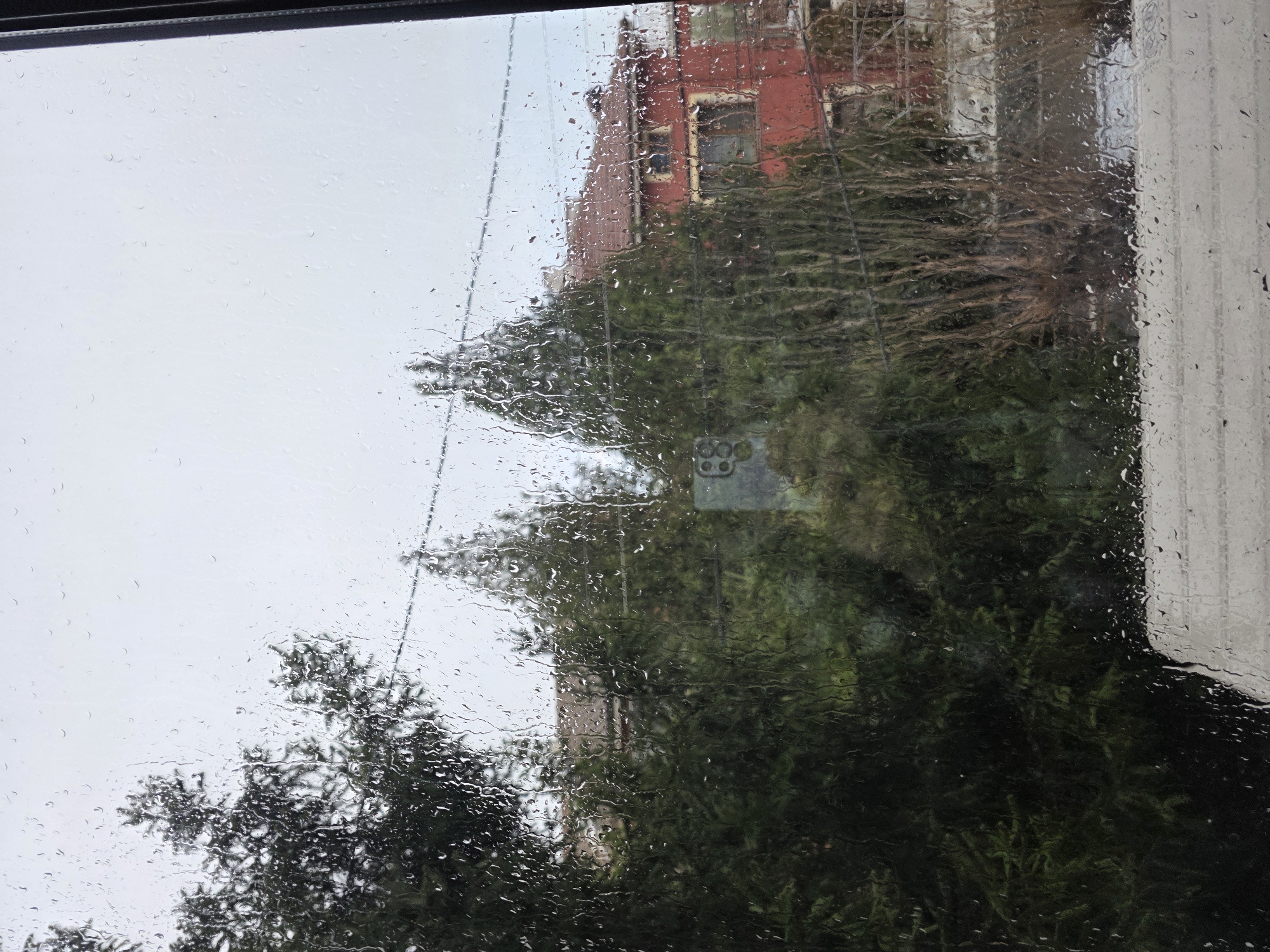 A view through a rain-streaked window. The foreground shows raindrops clinging to the glass, obscuring the scene outside. Visible through the window is a tall, likely multi-story red building with visible windows. Trees with dense green foliage partially obscure the building. The right edge of the frame shows a white vertical structure, possibly a wall or pillar of a building. The sky is overcast and gray. Specific location in Yagodovo, Bulgaria is indeterminate.