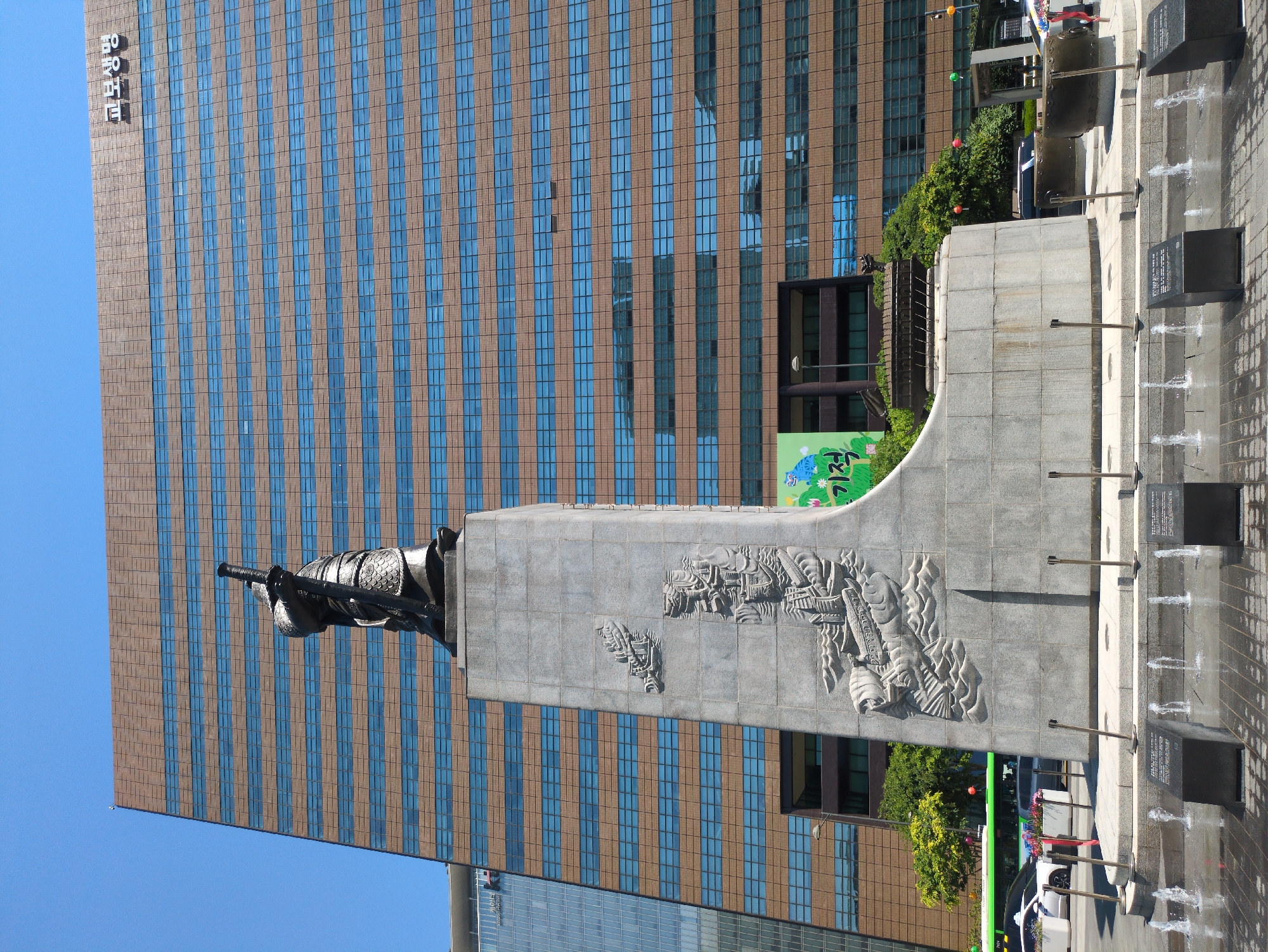 The image depicts a statue of Admiral Yi Sun-sin in Seoul, South Korea. The statue is mounted on a gray stone pedestal that has carved images on it. Behind the statue is a tall building with a grid pattern of brown and blue. A fountain is visible on the right side of the statue.
