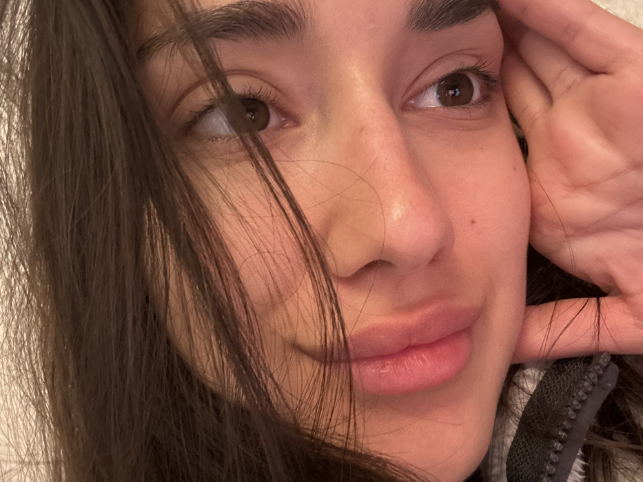 The image displays a close-up, angled portrait of a single female individual located in Zankovići, Montenegro. The subject has long, dark brown hair, some strands of which are draped across her face, partially obscuring her forehead and cheeks. Her skin tone is fair. Both of her eyes are visible, brown in color, with defined eyebrows and eyelashes. Her lips are full, appearing a natural pink, and are slightly relaxed. Her right hand, with fingers partially visible, supports the lower right side of her face near the jawline. A dark garment with a visible zipper, possibly a jacket or hoodie, is seen on the lower left side of the frame near her shoulder. The background is indistinct and appears to be a light, plain surface, suggesting an indoor environment. The lighting is soft and ambient.