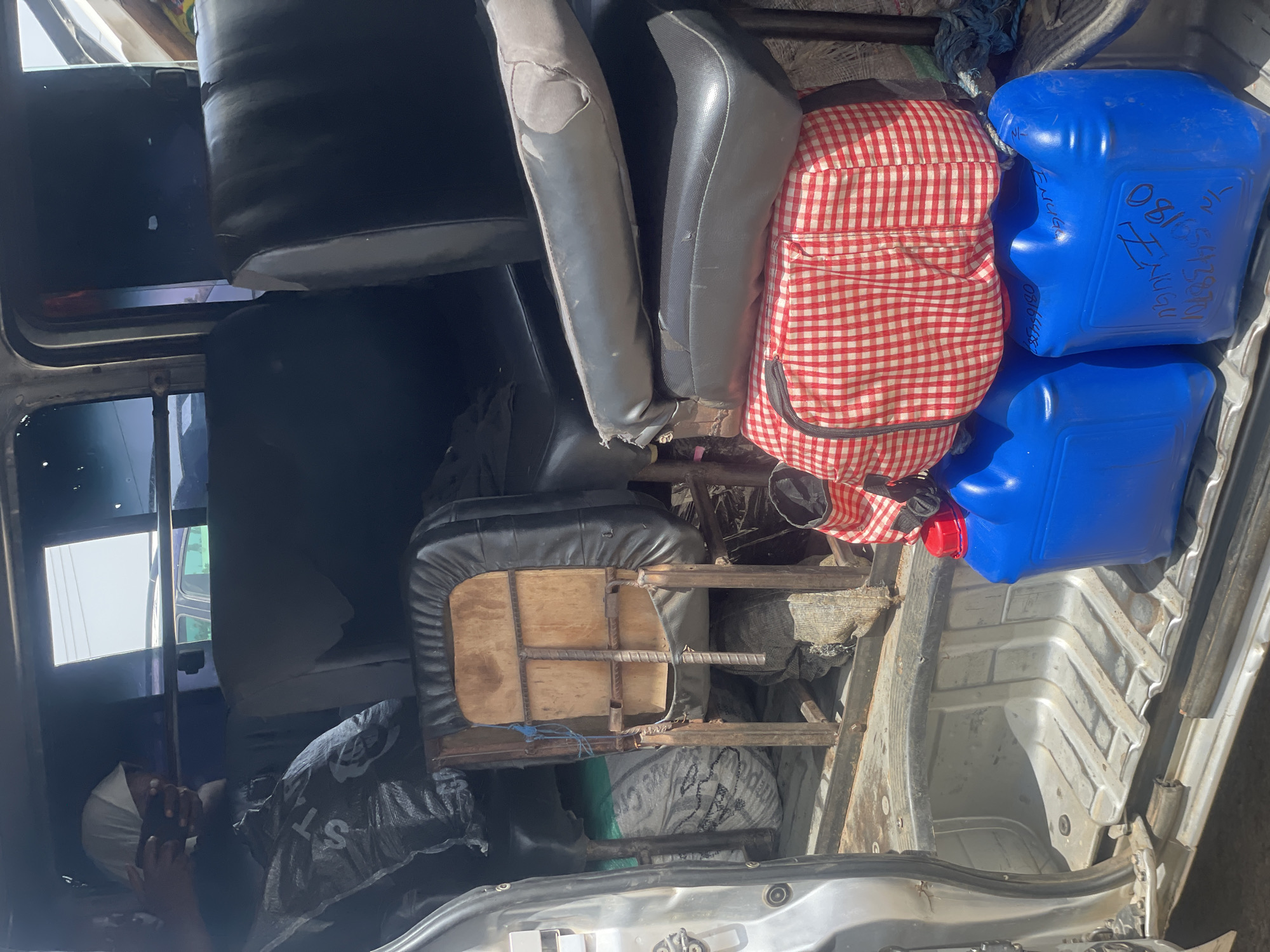 This image shows the interior of a vehicle, likely a van or truck, packed with various items. The focus is on the cargo area, which contains several blue plastic containers, possibly for water or fuel. One of these containers has writing on it in black marker, including what appears to be "081..." and "T/N NUGU". Next to the containers is a red and white checkered bag or backpack.
Further into the vehicle, there are several seats. One seat appears to be a simple metal frame with a wooden seat and some black upholstery. Another seat is a black padded seat, and further back, a gray seat is visible. A person, wearing a white head covering and a dark shirt, is lying down or resting in the lower part of the vehicle, partially obscured by black plastic bags and other items.
The scene appears to be outdoors, with sunlight casting shadows and highlighting the metal interior of the vehicle. The overall impression is one of a load being transported, possibly for distribution or personal use, given the presence of the containers and the person resting among the cargo. The setting in Bauchi, Nigeria, suggests a context of everyday life and transportation of goods in that region. There are no obvious cues about the time of day or weather, beyond the presence of sunlight.