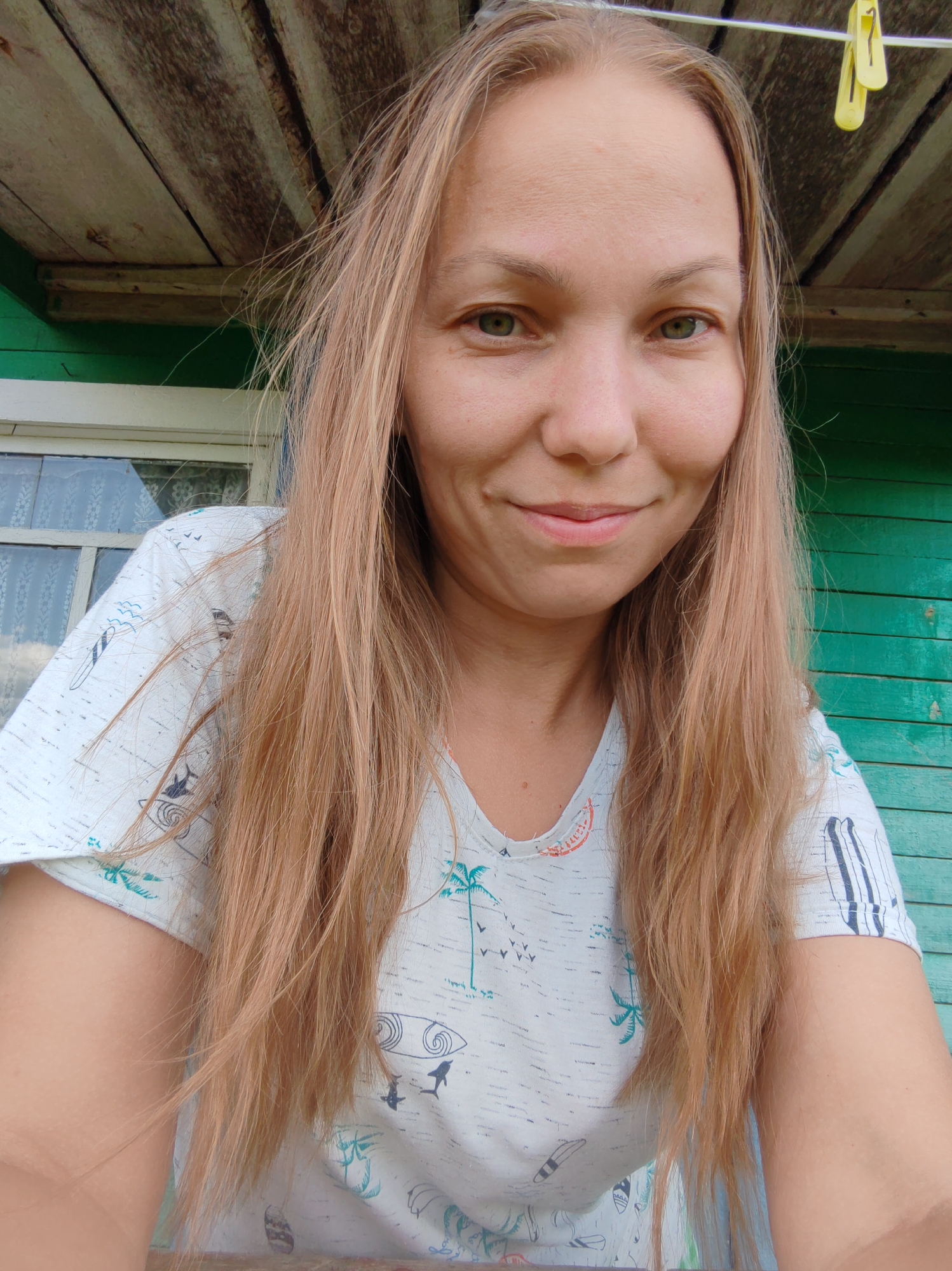 A medium-shot, front-facing selfie of a woman with long, light brown hair and green eyes is taken outdoors. She is smiling slightly, with a hint of a blush on her cheeks. She is wearing a light-colored t-shirt with a pattern of palm trees and surfboards. The background features a green wooden wall and a wooden ceiling with visible planks. A yellow clothespin is clipped to a wire above her head. The scene appears to be in an outdoor, rustic setting, possibly a porch or shed, given the wooden structures and the rustic aesthetic. The location is indicated as Чухлэм, Russia.