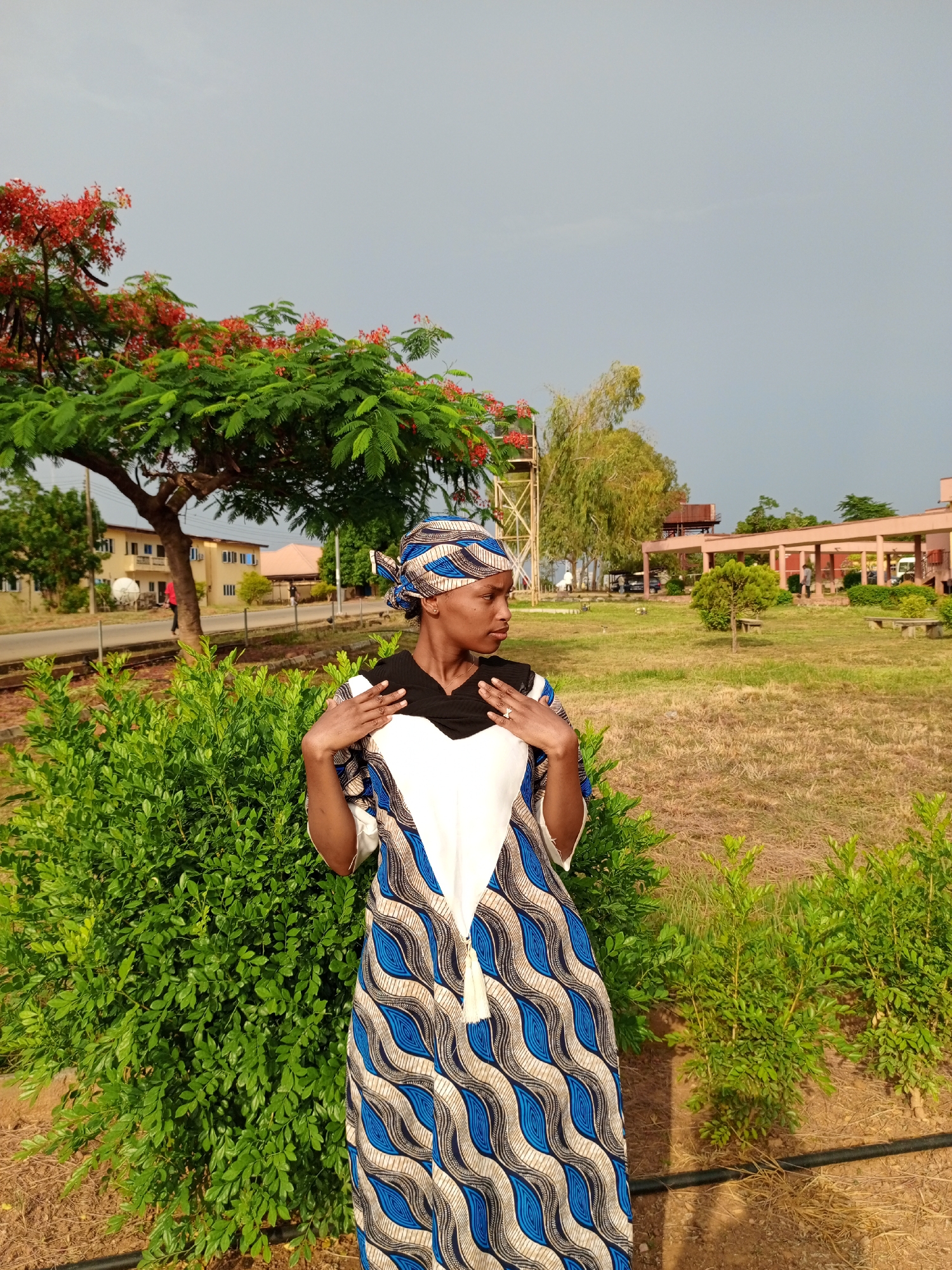 A woman stands outdoors next to a flowering tree and a green bush in Keffi, Nigeria. She is wearing a blue, white, and black patterned dress and a matching headscarf. The woman has her hands up near her chest and is looking to the side. In the background is a grassy lawn with trees and buildings, under a cloudy sky. A water tower is visible in the background.