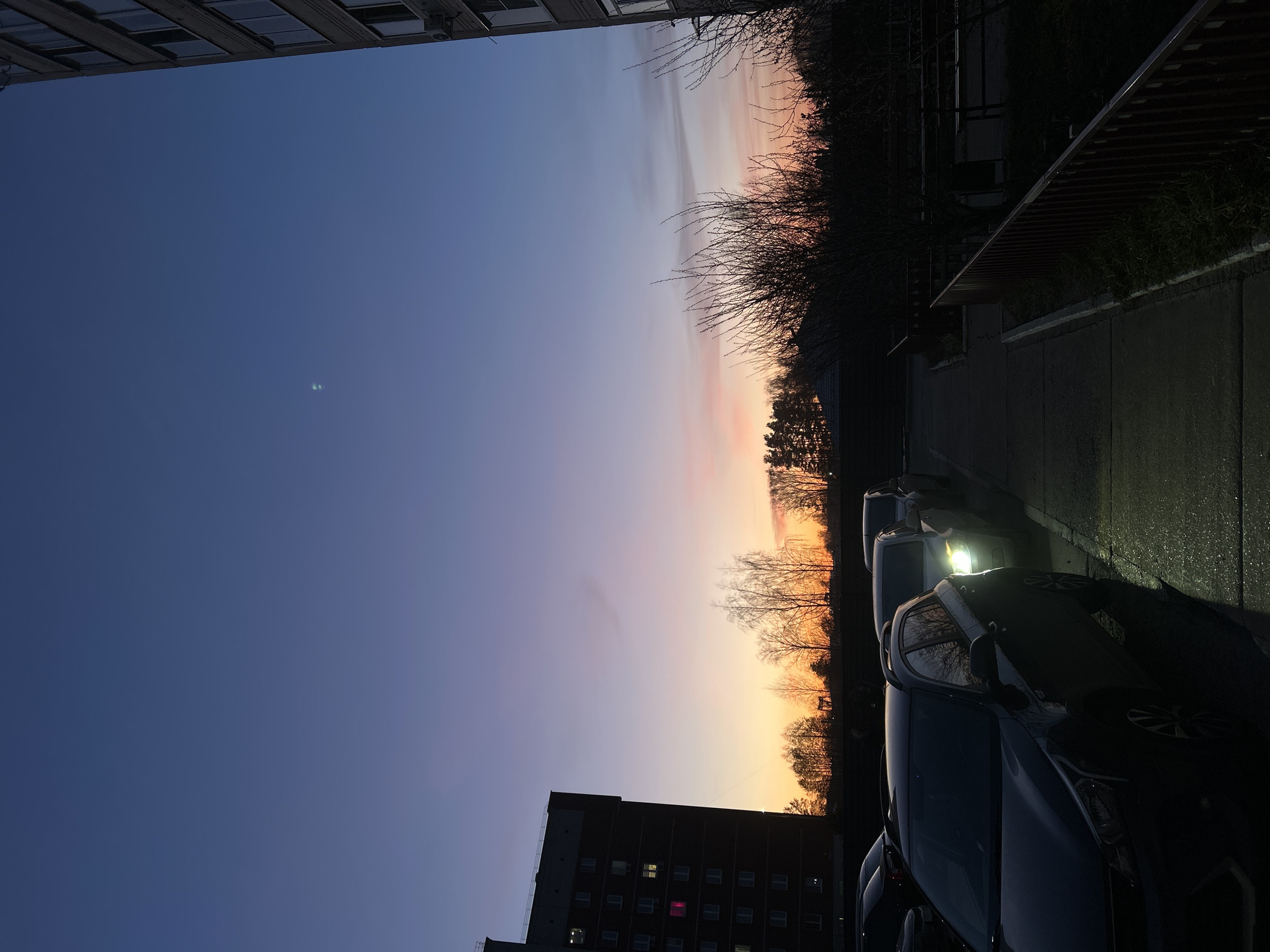 The image captures a serene urban scene at sunset or dusk in Chaikovsky, Russia. The sky, dominating the upper portion, displays a beautiful gradient of colors, transitioning from a deep indigo blue in the upper left, where a faint crescent moon is visible, to soft purples, pinks, and vibrant oranges along the horizon on the right.
Silhouetted against the colorful sky are the bare branches of numerous trees and bushes, suggesting late autumn or winter. In the upper left, a portion of a multi-story building, likely an apartment complex, is visible. In the lower left, another dark, multi-story building with several illuminated windows, one distinctly glowing pink, adds artificial light to the darkening scene.
The lower right half of the image features a paved surface, possibly a street or parking lot, which appears wet and reflective. Several cars are parked along this surface, their dark forms contrasting with the fading light. One car in the mid-right foreground has its bright LED headlights on, casting a distinct glow on the wet pavement. Adjacent to the parked cars, a dark, angular structure with horizontal slats and some green foliage can be seen, possibly a covered walkway or a architectural feature. No explicit activity is taking place, and no visible text is present.