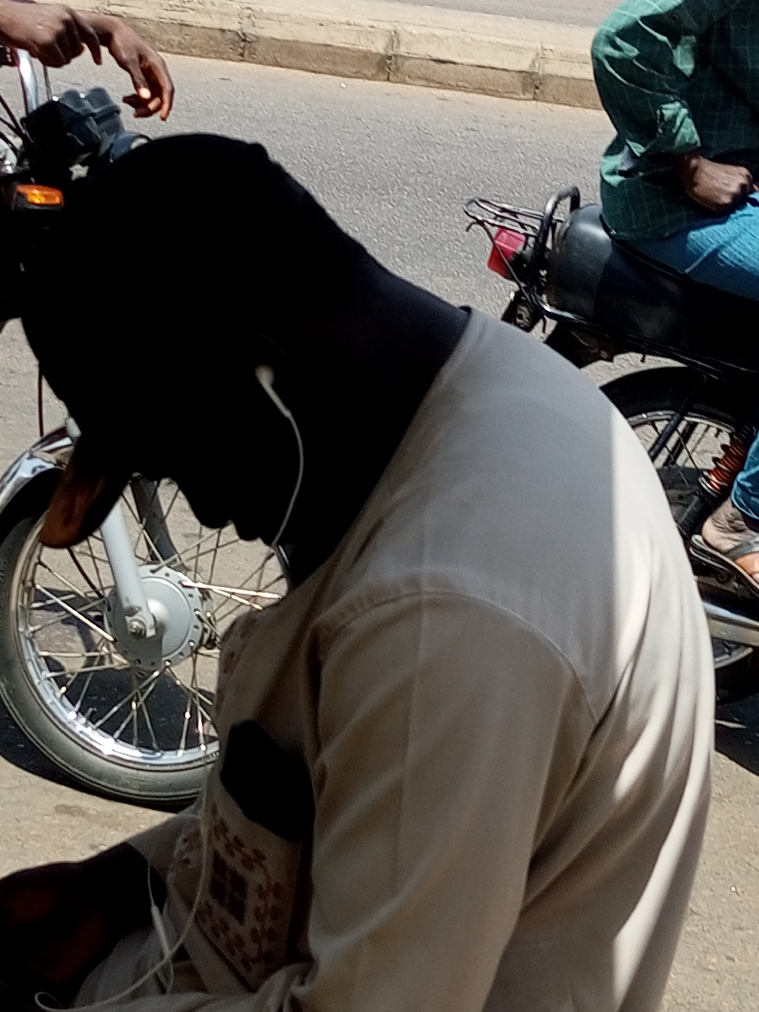 In the city of Gombe, Nigeria, a person is seated on a motorcycle, their body angled to the right. They are wearing a light-colored shirt and have earbuds in their ears. The motorcycle's front wheel, fender, and handlebars are visible, with a chrome fender and a spoke wheel. To the right, another person is seated on the same motorcycle, wearing a green checkered shirt and blue pants. Their legs are visible, and they are wearing sandals. The motorcycle has a black seat, a rear rack with a red taillight, and suspension components. The scene takes place on a paved road with a concrete curb. Another person's hand is visible on the left, appearing to be holding the motorcycle's handlebars.