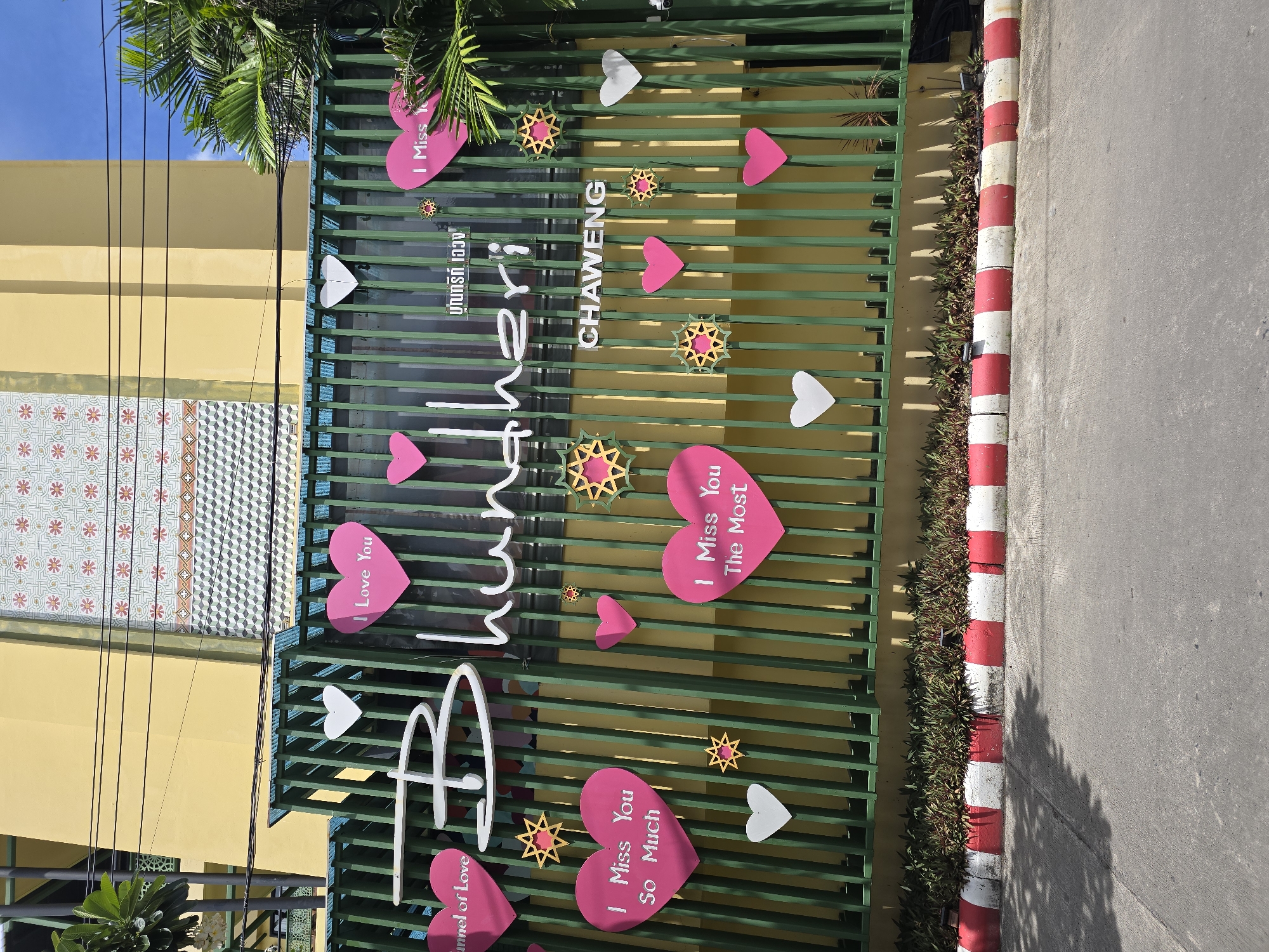 The image shows a green metal fence in front of a yellow building. The fence is decorated with various pink and white hearts, as well as some star-shaped ornaments. Some of the hearts have text written on them. The building behind the fence has a patterned facade and a blue sky with clouds is visible above. There are also some palm trees on the right side of the image. A red and white striped curb runs along the bottom of the image, separating the fence from a paved surface, likely a road. The scene appears to be outdoors in a sunny environment.