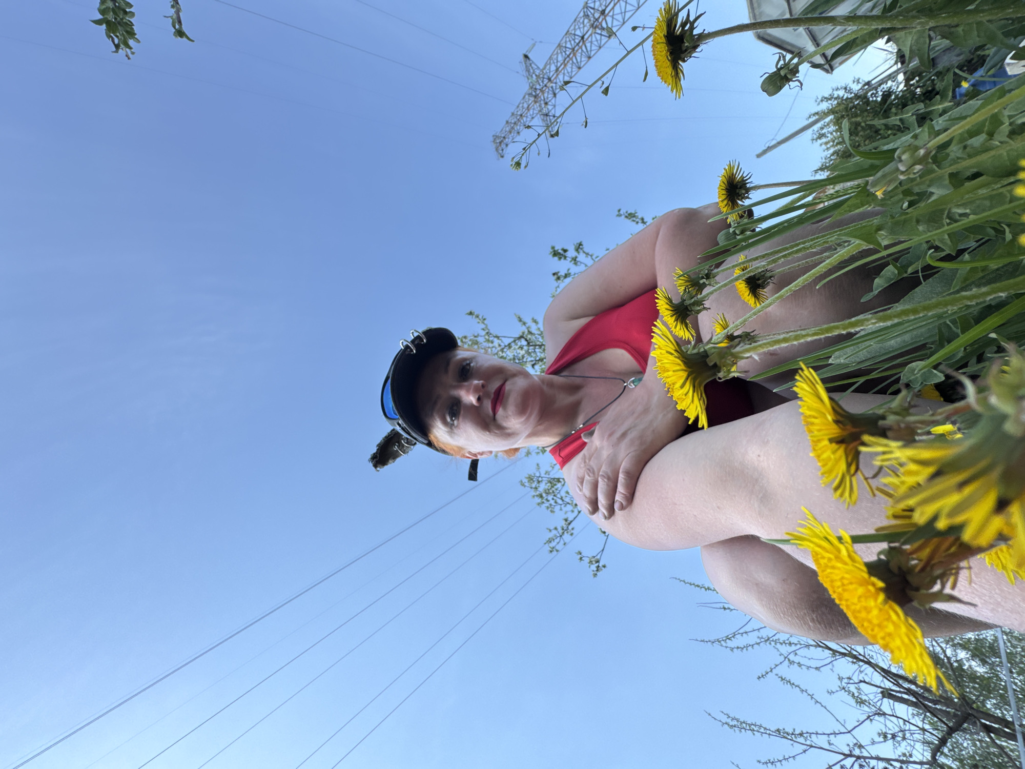 A low-angle shot depicts a woman outdoors in Chaikovsky, Russia. She is wearing a red top, a cap with goggles, and a necklace. Dandelions are prominently featured around her. Overhead power lines and a transmission tower are visible against a clear blue sky.