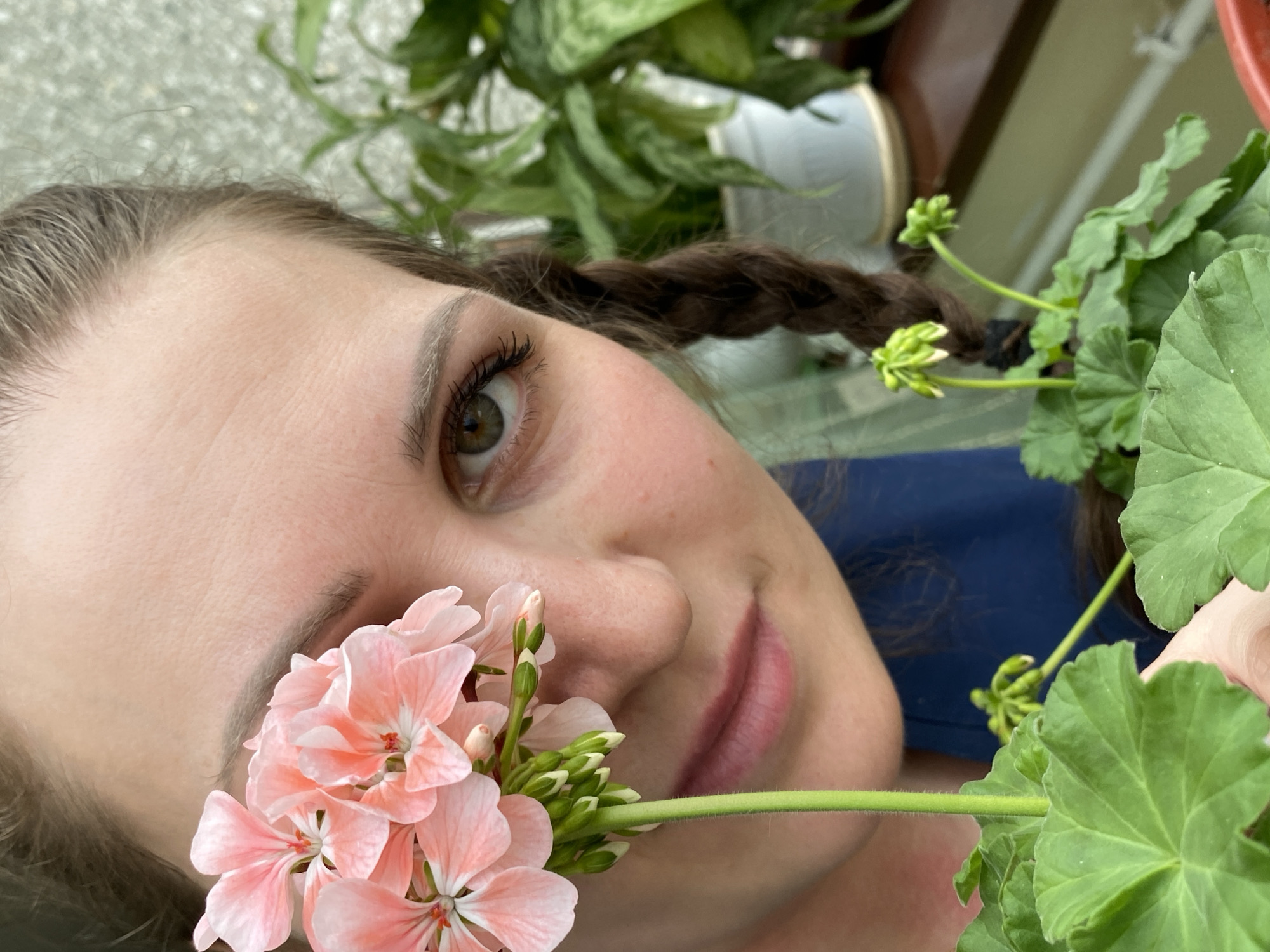 A close-up, slightly tilted, shot features a fair-skinned woman with braided brown hair, posed next to a flowering plant. The pink flowers are near her left eye and cheek. Her eye color appears to be hazel. Green leaves and flower buds surround her face, along with another potted plant in the background. A section of grey surface is visible in the top-left corner. The woman is wearing a blue top. There is no information to determine if this image was taken in the city of Марковский, Russia.