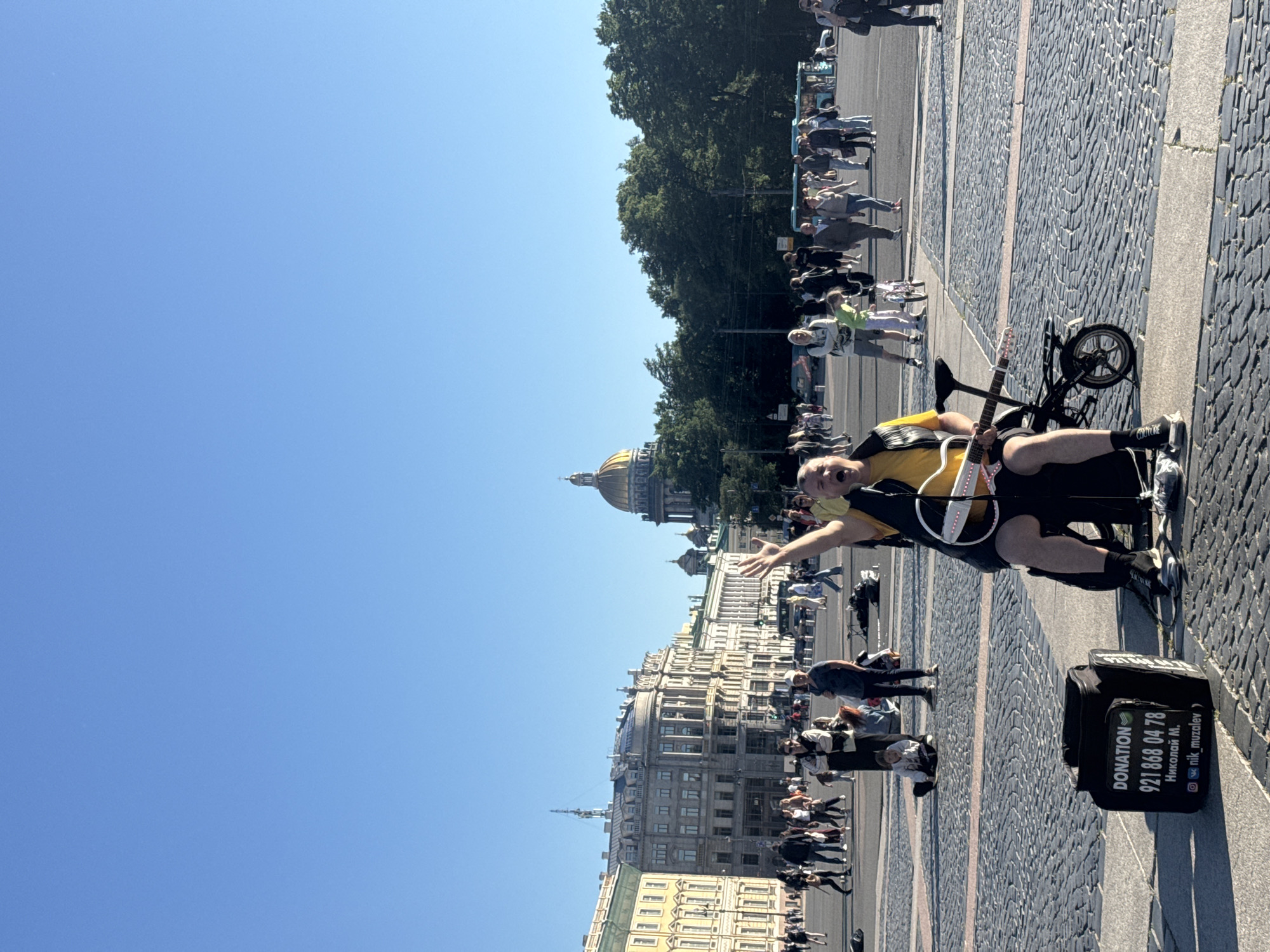 A man is playing an electric guitar in Palace Square, Saint Petersburg, Russia. He is seated on a black bag which is labeled "DONATION" with a phone number and social media handle. He wears a yellow shirt and a black vest, with black shorts and sneakers. His right arm is raised. The guitar has white and pink lights along its neck. In the background, numerous people are walking and standing around the square. The architecture of buildings in the Russian Imperial style is visible, including the golden dome of Saint Isaac's Cathedral. The sky is clear and blue. The ground is paved with cobblestones.