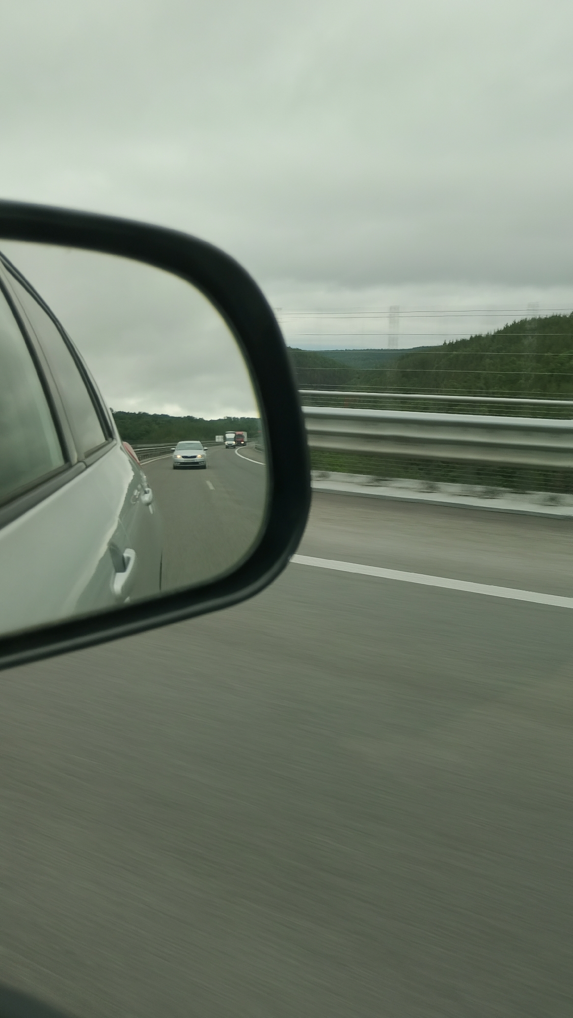 The image is a side-view mirror reflection of a highway in Baliovtsi, Bulgaria. In the reflection, a silver car is visible in the distance, followed by a red truck. The highway has white lane markings and a metal guardrail on the right side. Beyond the highway, there are green trees and a cloudy sky. The overall scene suggests a drive on a cloudy day.