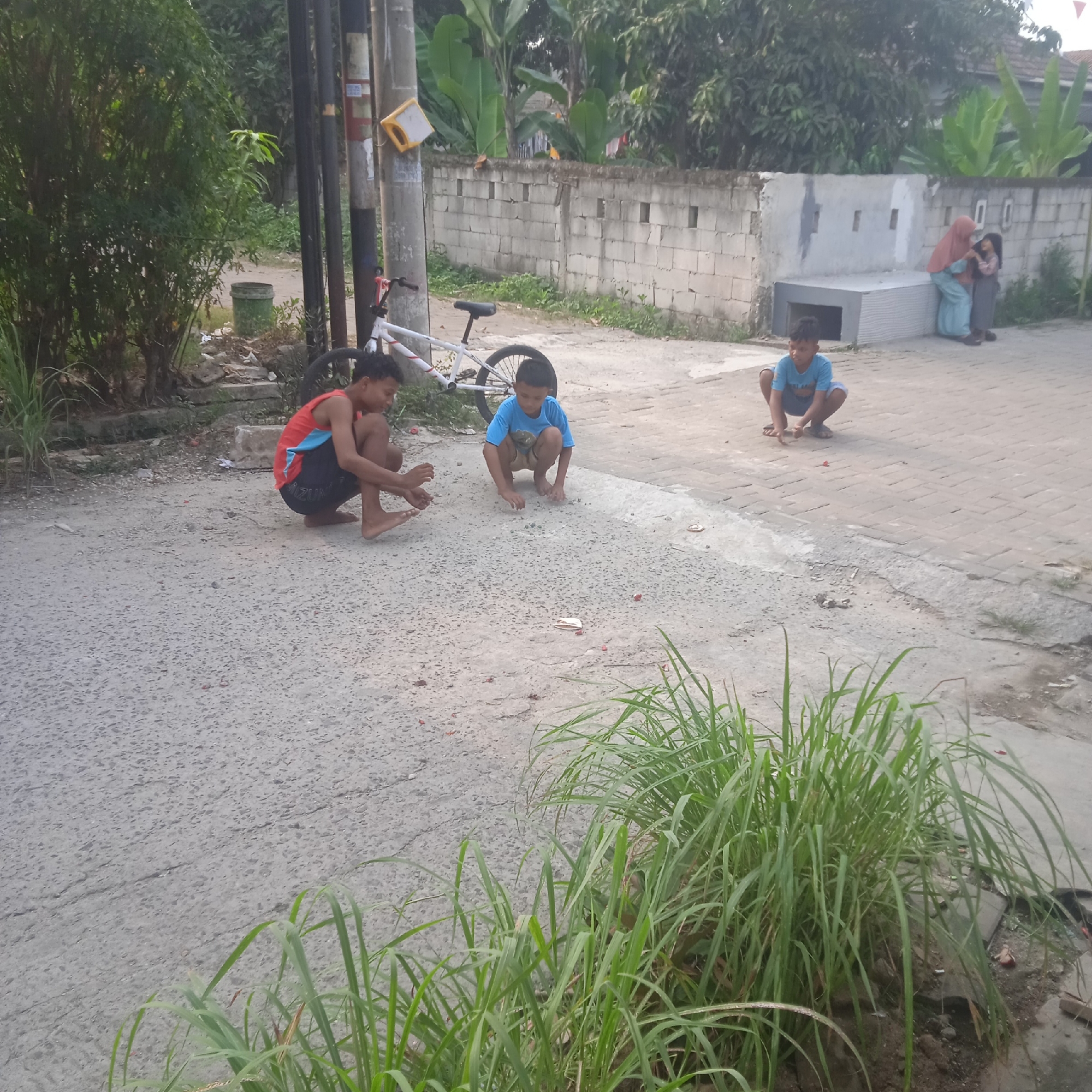 The image captures a street scene in Situterate, Indonesia, with three young boys engaged in play. Two boys are crouched on the asphalt roadway, while a third is seated on the paved sidewalk. A white bicycle with red accents is parked on the left side of the road. The background features a low concrete block wall, vegetation, and a building. A woman and a girl are visible in the distance, near the wall, with the woman appearing to interact with the child. The ground is littered with small objects, possibly debris or game pieces. The lighting suggests it is daytime.