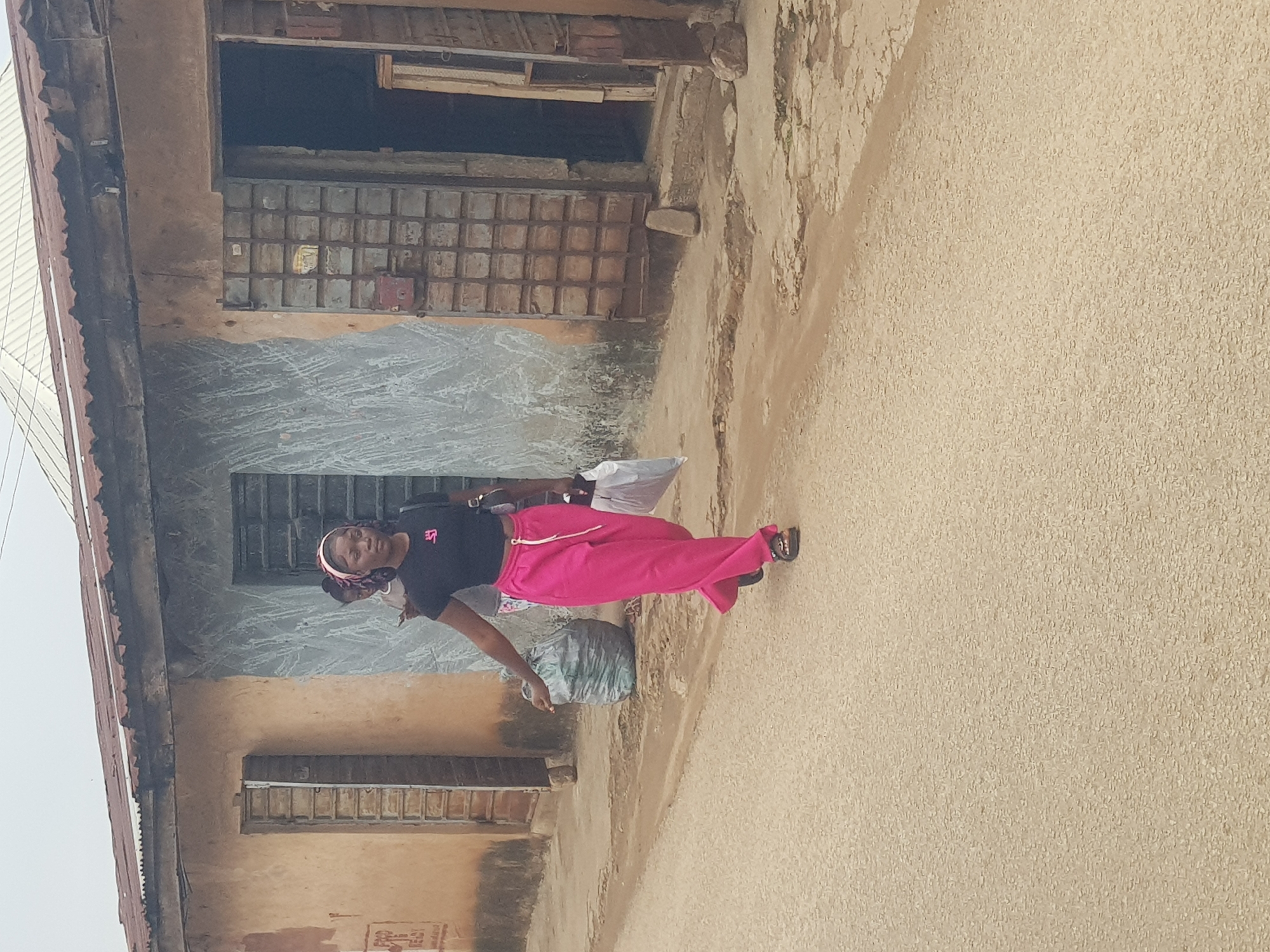 A young woman walks on a dusty street in Jos, Nigeria, carrying a white plastic bag and a large blue garbage bag. She wears a black crop top and bright pink pants, with a pink headscarf. In the background, a dilapidated building shows signs of decay, with a corrugated iron roof and a weathered facade. The street is unpaved, with visible potholes and debris. Another person, partially obscured, is visible behind the young woman. The overall scene suggests a working-class or informal settlement.