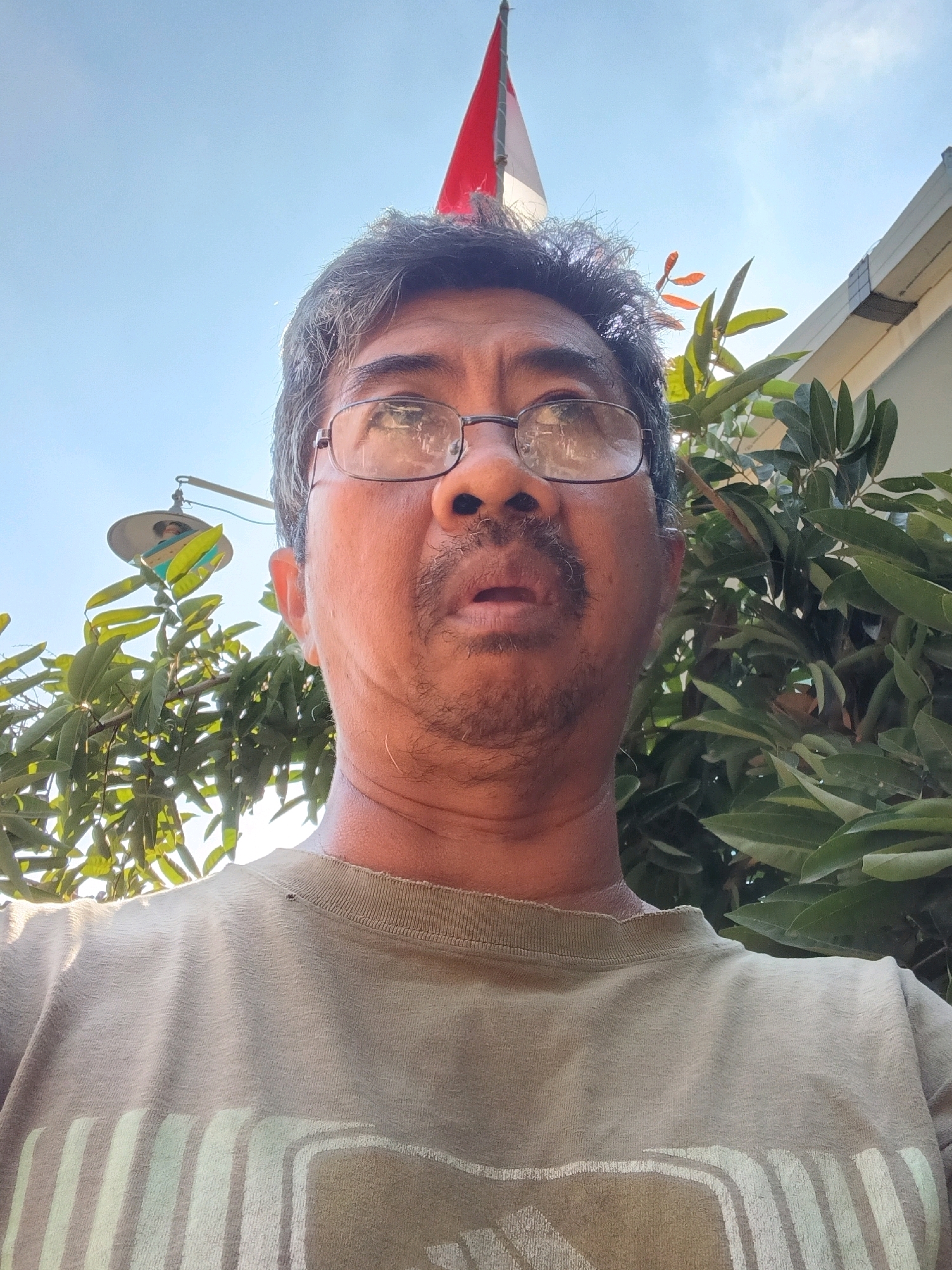A low-angle, close-up selfie of a middle-aged man with glasses is taken outdoors. The man has graying hair, a mustache, and is wearing a tan t-shirt with a striped design. He is looking directly at the camera with his mouth slightly open. In the background, there is a blue sky, a red and white flag, green foliage, and a portion of a white building. A light fixture with a decorative shade is also visible hanging from a pole among the trees. The image appears to be taken in Tanggul Wetan, Indonesia, suggested by the presence of a flag and the common appearance of the architecture and vegetation in that region. The overall lighting suggests it is daytime, likely sunny, with the sun casting highlights on the man's glasses and hair.