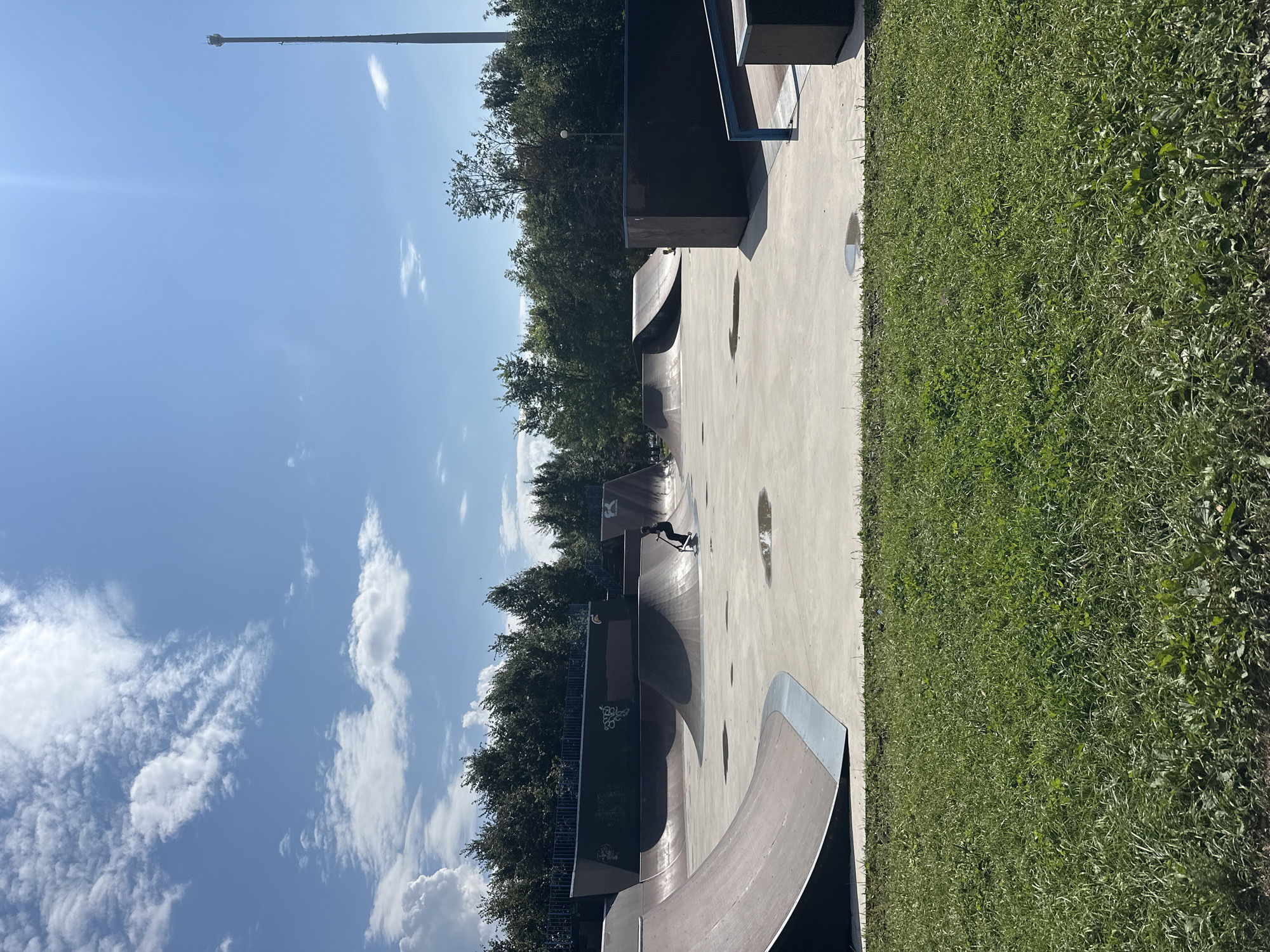 The image depicts an outdoor skatepark located in Saint Petersburg, Russia. The skatepark features a concrete surface with various ramps, bowls, and ledges, constructed from a combination of concrete and metal. In the mid-ground, a single individual is seen on a scooter, actively engaged in riding within the park. The surrounding environment includes a lush green lawn in the foreground and a dense line of trees in the background, suggesting a park or recreational area. The sky above is bright blue with scattered white clouds, indicative of fair weather. A tall communication tower is visible in the distance on the right side of the frame. The overall scene captures a moment of recreational activity in a public space.