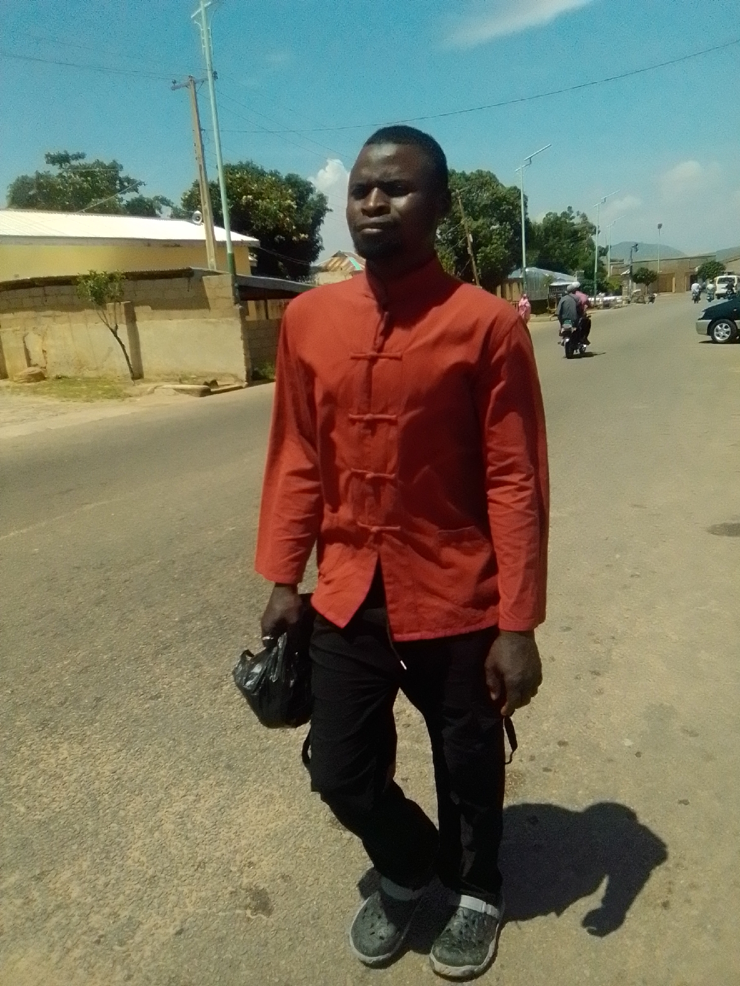 A man is standing in the middle of a paved road in the city of Galadima, Nigeria. He is wearing a red, mandarin-collar shirt with frog closures and black pants. He is holding a black plastic bag in his left hand. His shadow is cast on the road beside him. In the background, there are buildings, trees, a street light, and other people and vehicles. The sky is blue with some clouds.