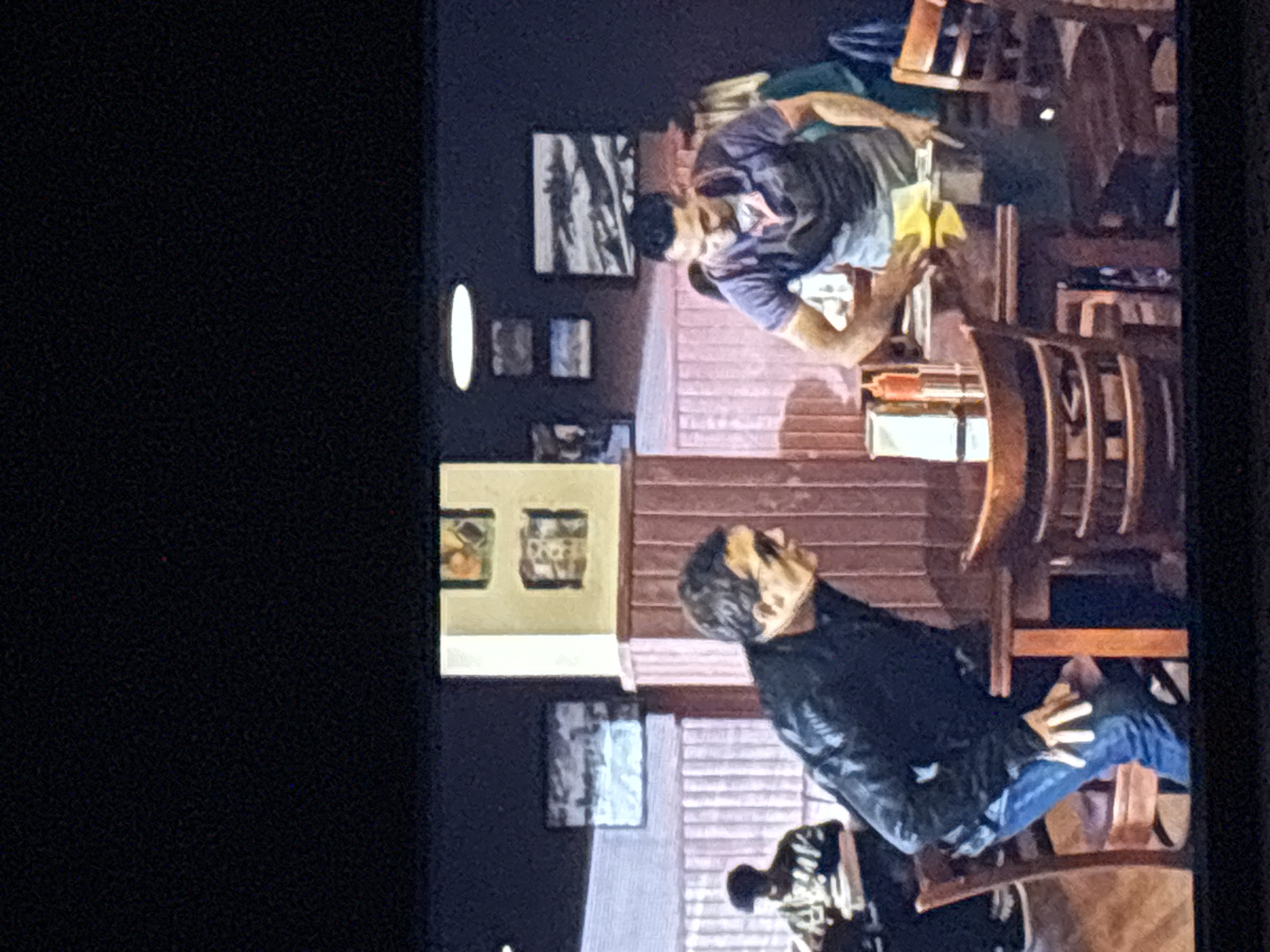 This image depicts an indoor scene, likely a restaurant or cafe, featuring two men. The photograph is oriented vertically, rotated 90 degrees clockwise.
In the foreground, positioned on the right side of the frame (when viewed correctly), a man is seated. He wears a dark, possibly black, jacket and blue jeans, with his left hand resting on his knee. He has dark hair and appears to be looking upwards and slightly to his left, seemingly observing something or someone beyond the frame.
Further back, towards the left of the frame, another man is seated at a wooden table. He is wearing a short-sleeved, grey or purple t-shirt and has dark hair and a beard. His gaze is directed downwards towards the table, where he seems to be handling a small, yellow object or cloth.
The setting is furnished with multiple wooden tables and chairs, characteristic of a dining establishment. The walls are adorned with various framed pictures. A distinctive white oval light fixture is visible on the dark wall in the background. The wall behind the tables features reddish-brown horizontal paneling. A light-colored tablecloth or napkin is visible on the table where the man in the t-shirt is seated. The lighting suggests an evening or nighttime atmosphere. No visible text is discernible within the image. The location context for this scene is Budva, Montenegro.