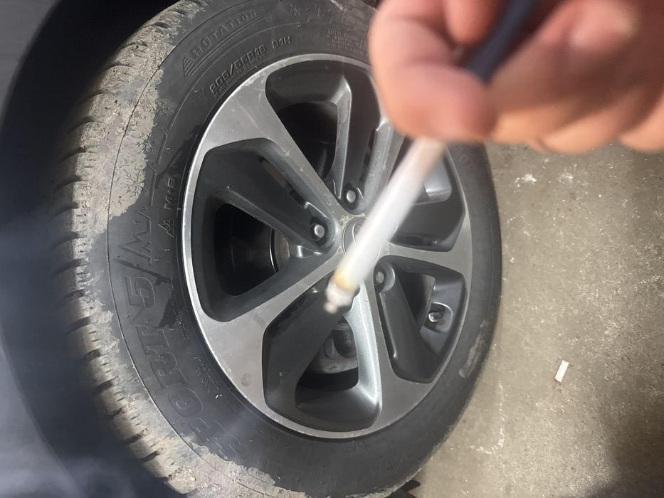 The scene is situated in Maslarevo, Bulgaria. The image presents a close-up, low-angle perspective focusing on a lit cigarette held by a human hand in the foreground, with a vehicle's front passenger side wheel as the primary background subject.
The ground appears to be a concrete or asphalt surface, scattered with light dust or debris. A distinct white cigarette filter or butt lies on this surface beneath the car wheel, towards the lower-left.
A right human hand, with fingers partially visible, holds a lit cigarette diagonally across the mid-frame. The cigarette's tip is amber, indicating it is actively burning, and a thin stream of white smoke emanates from it, rising towards the upper-left where it dissipates into a larger, hazy plume.
The car wheel consists of a black rubber tire and a five-spoke alloy rim. The tire, identified as "SPORT 5 M+S," displays markings including "RADIAL TUBELESS," a "ROTATION" arrow, and size specifications "205/55R16 91H." Multiple white snowflake symbols are visible on the tire's sidewall, indicating its winter classification. The tire's surface, particularly the tread and portions of the sidewall, is coated with dried mud or dirt. The alloy rim features a two-tone design with dark grey/anthracite inner spokes and silver/machined outer edges, secured by five silver lug nuts. A brake disc is partially visible behind the rim spokes.