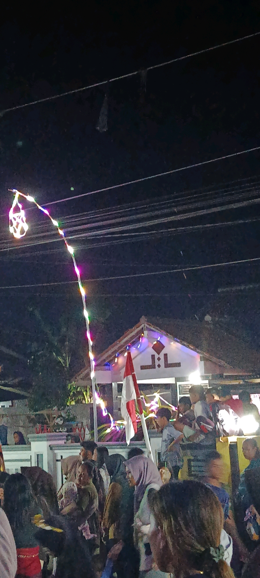 This image captures a nighttime outdoor gathering in Binangun, Indonesia. A string of colorful LED lights hangs across the scene, suspended by wires above a group of people. The lights trail down from a decorative lantern on the left, illuminating a pole with an Indonesian flag attached. In the background, a white building with a tiled roof is visible, adorned with more lights and a symbol resembling "JL:L". A crowd of individuals, including children and adults, are gathered in front of the building, some holding babies. The atmosphere appears festive, suggested by the lighting and the presence of a flag, likely indicating a local celebration or event. The overall setting is a residential street or common area at night.