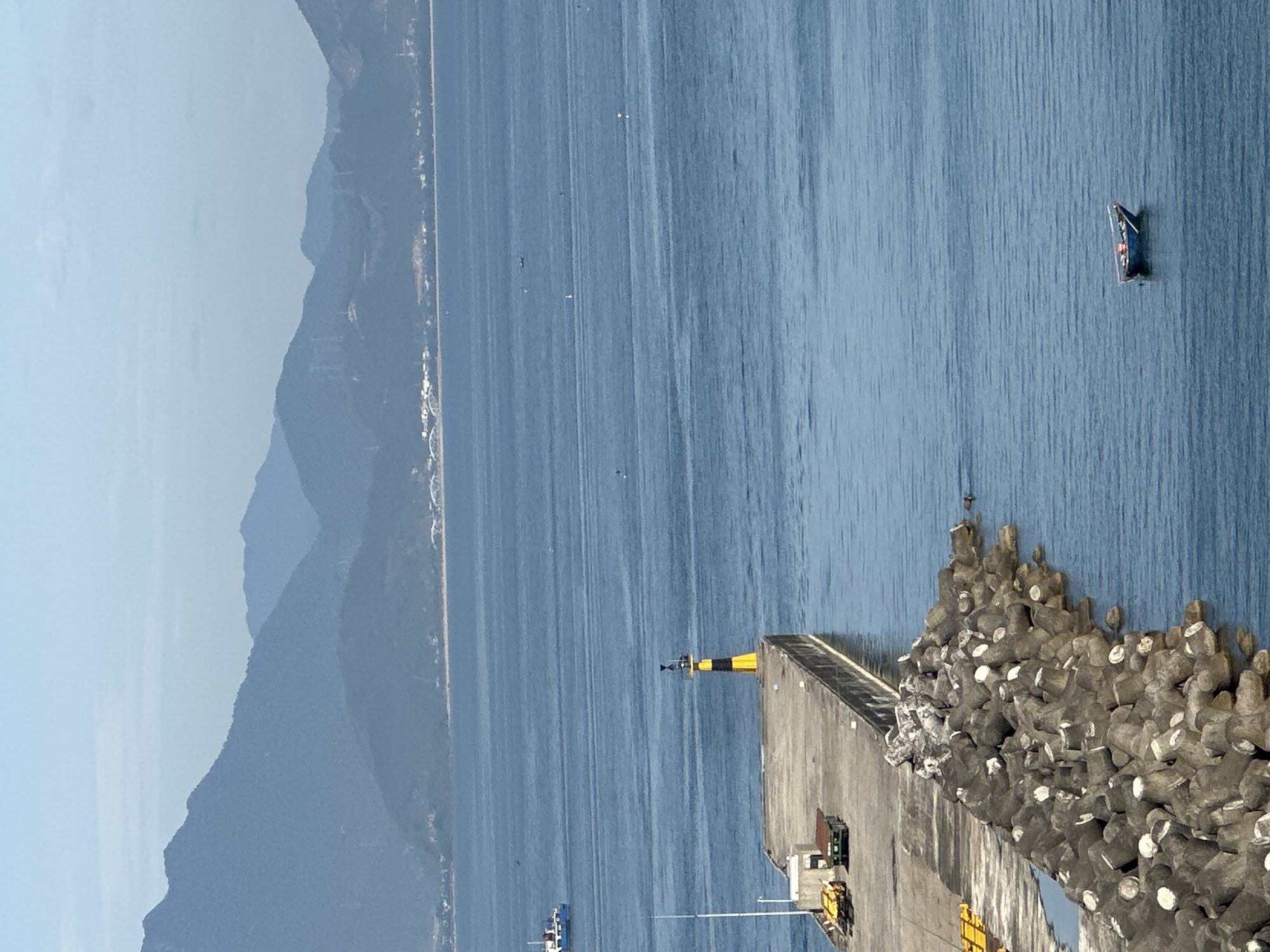 The image depicts a tranquil seascape with a prominent pier in the foreground. The pier is constructed of concrete and lined with large, angular concrete blocks forming a breakwater. On the pier, a yellow and black lighthouse stands tall, while two yellow forklifts and a dark green cart are parked nearby. A narrow, dark blue boat is visible in the distance, adrift on the calm blue water. The background is dominated by a range of hazy blue mountains, suggesting a coastal mountainous landscape. In the far distance, a white building with a bridge is visible along the coastline, and a blue and white ship sails on the horizon. The overall scene is peaceful and scenic, hinting at a location such as the Son Tra Peninsula in Da Nang, Vietnam, known for its natural beauty and coastal features.