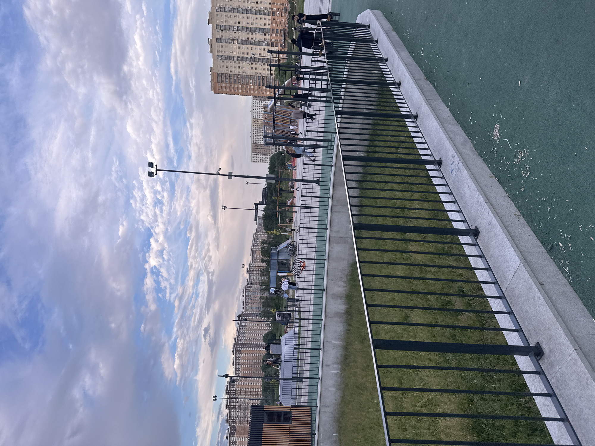 The image depicts a public park in Moscow, Russia, featuring a modern outdoor sports and recreation area. In the foreground, a dark metal fence lines a paved green surface, with a concrete ledge below. Beyond the fence, a grassy area slopes downwards. In the midground, a children's playground with slides and climbing structures is visible, along with an area for calisthenics. Several individuals are scattered throughout the park, some engaged in activities and others walking. In the background, a row of tall, multi-story residential buildings characteristic of Moscow's urban landscape stretches across the horizon. The sky is partly cloudy, with a mix of blue sky and white clouds, indicating daytime. The overall scene suggests a well-maintained urban park designed for community recreation.