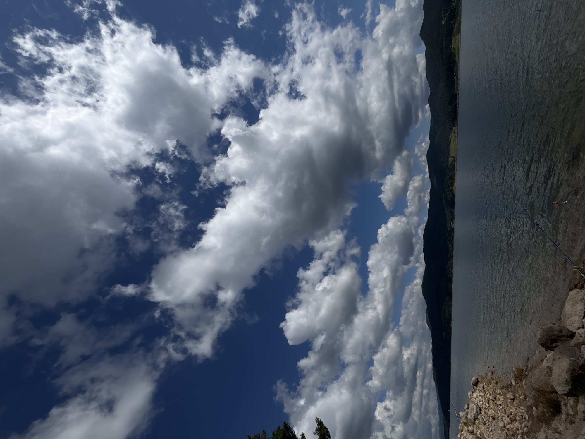 An outdoor, vertical, eye-level shot captures a scenic view of a vast, calm body of water under a dramatic sky. The water occupies the majority of the frame, reflecting the sky above and the dark, tree-covered mountains that form the horizon. The shoreline in the foreground is rocky and uneven, with a few larger boulders. A single fishing rod with a red float is visible in the water, suggesting activity. The sky is a vibrant blue, populated by a mix of fluffy white cumulus clouds and darker, more imposing storm clouds, creating a dynamic and textured canopy. The overall impression is one of natural beauty and tranquility.