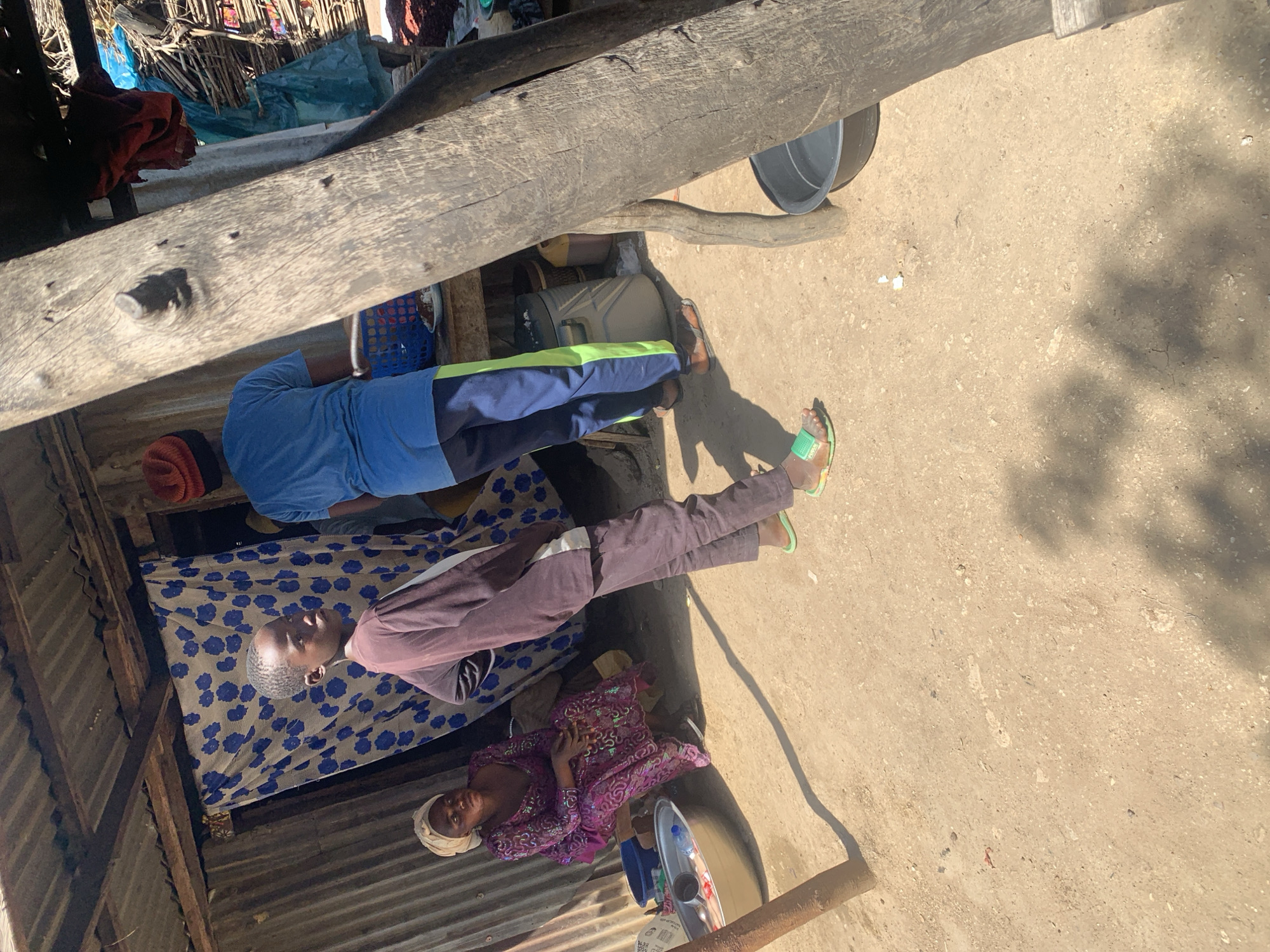 In Project, Nigeria, a scene unfolds under a makeshift, corrugated metal roof supported by rough-hewn wooden beams. The ground is a mixture of compacted dirt and sand. In the mid-ground, a young person with short dark hair, wearing a dark brown long-sleeved shirt and dark pants, stands facing left, looking towards the camera. They are wearing light green flip-flops. To their left, a woman in a vibrant purple, intricately patterned dress and a light-colored headscarf sits on a low wooden bench. Before her, a large metal basin contains water and a bottle. Behind the young person, another individual, partially obscured, wears a bright blue t-shirt and dark pants with a green stripe down the side, along with a red beanie. They are looking towards a blue plastic basket on a wooden table. To the far right, a thick, weathered wooden post dominates the foreground, casting a strong shadow. On the ground near this post is a dark basin filled with water. The background reveals more of the rustic shelter with draped fabric and various items, suggesting a functional, outdoor living or working space.