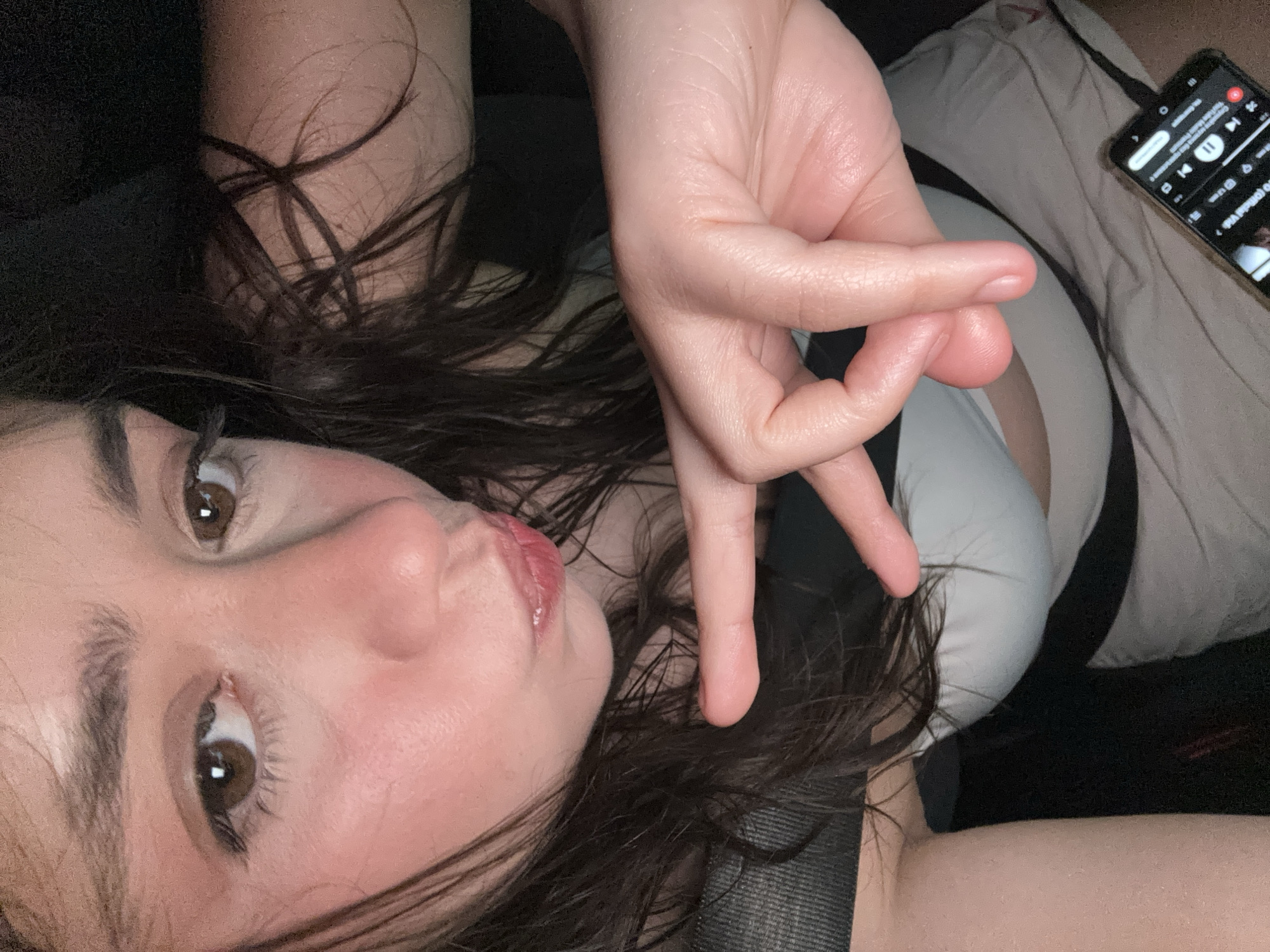 A woman with brown hair and eyes is visible from the chest up, angled to the left. She is wearing a white top and a black seatbelt, and making a hand gesture with her right hand. A smartphone with a music player open is visible to the right of her. The location is likely inside a car. There are no indicators of Koprivlen, Bulgaria.