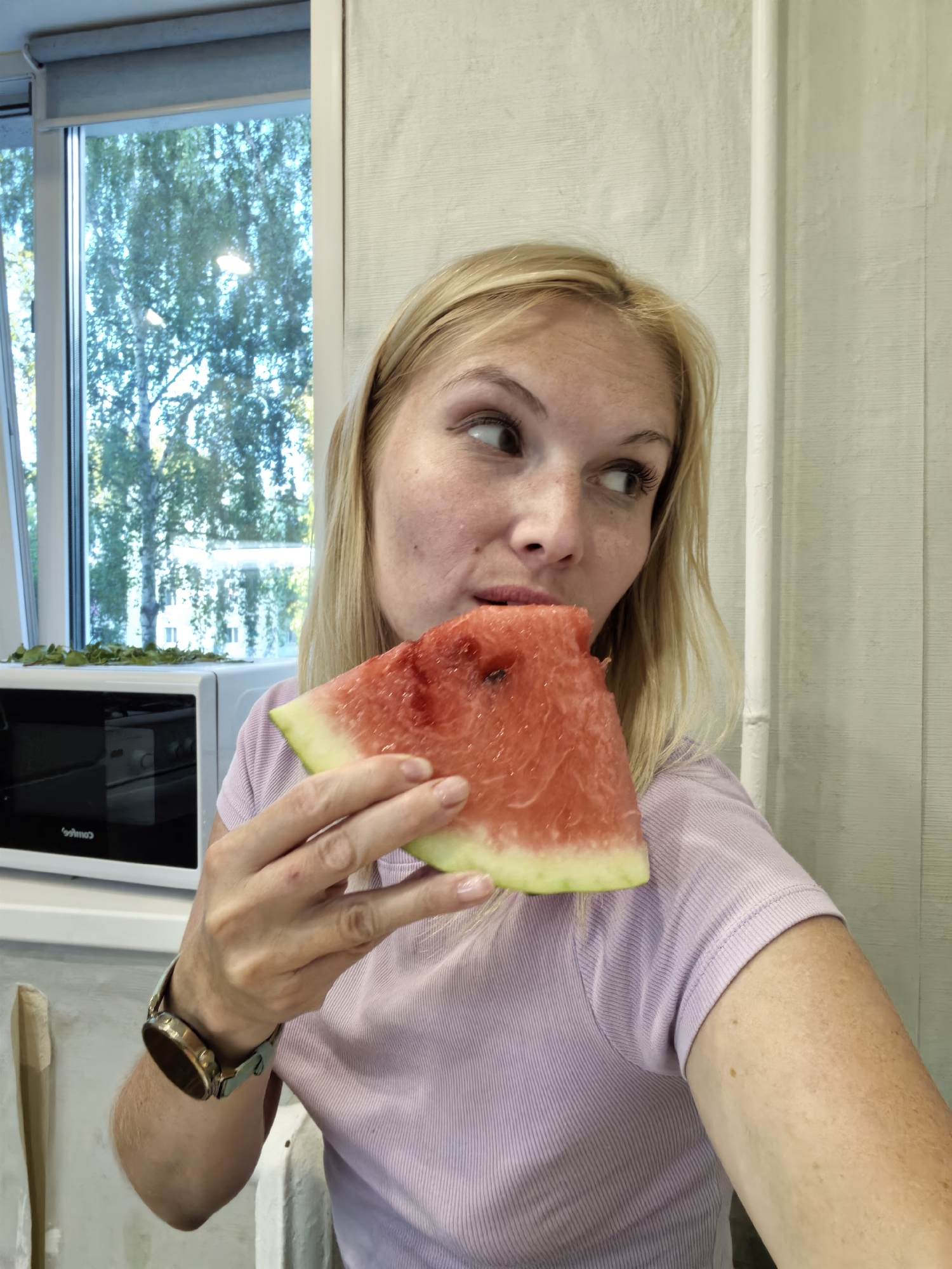 A blonde woman is eating a slice of watermelon in what appears to be a kitchen. She is wearing a light purple t-shirt and a watch on her left wrist. Behind her, there is a microwave and a window looking out onto trees. The scene is brightly lit, with natural light coming from the window. The overall impression is that of a casual, everyday moment.