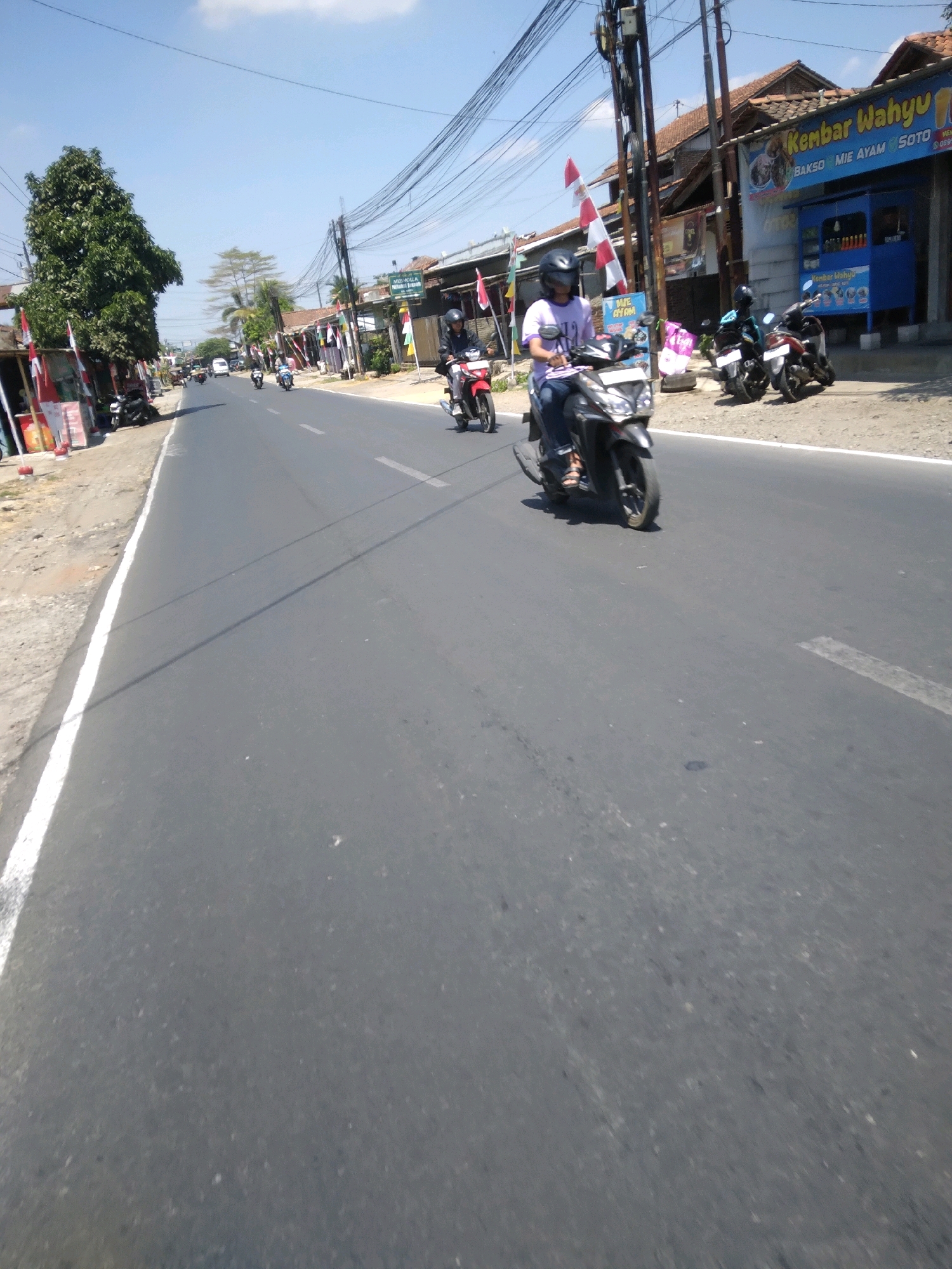 The image depicts a street scene in Sokaraja, Indonesia, on a sunny day with a clear blue sky. A paved road stretches into the distance, bordered by a dirt shoulder and various buildings on the right side. The buildings appear to be shops and residences, with awnings and signage visible. One sign reads "Kembar Wahyu" and advertises "Bakso Mie Ayam Soto". A blue kiosk is situated in front of this establishment.
Several motorcycles are present on the road. In the foreground, a person wearing a helmet and a light purple shirt is riding a dark-colored scooter. Behind them, another motorcycle is visible. Further down the road, two more motorcycles are seen, one with a rider wearing a blue shirt and helmet, and another dark-colored motorcycle. On the left side of the road, there are trees and more buildings, some decorated with Indonesian flags. Power lines are strung across the scene, supported by poles. The overall impression is a busy, sunlit street in a commercial or residential area of Sokaraja.