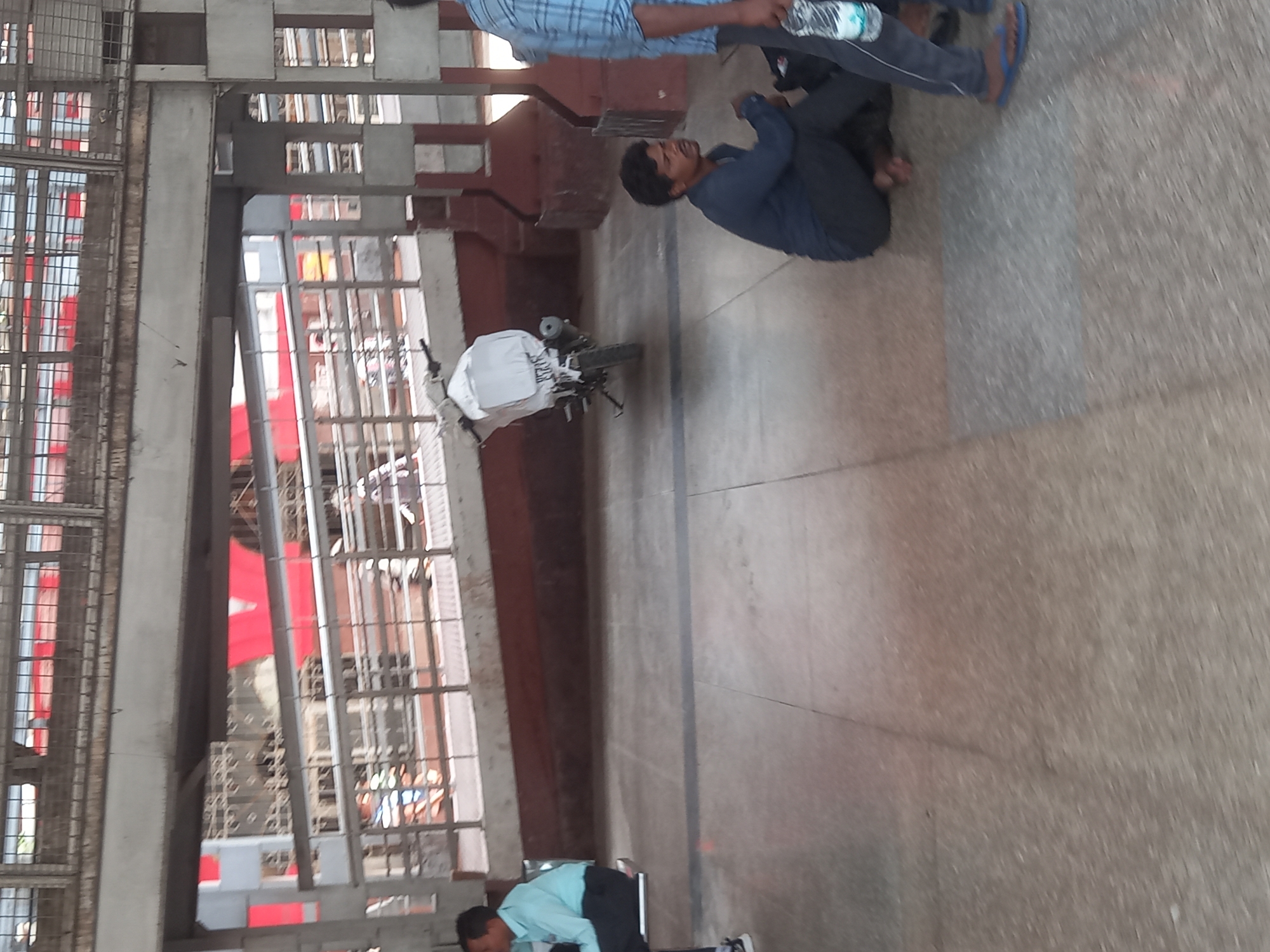 The image depicts a scene at the Jhansi railway station in Jhansi, India. The setting appears to be an outdoor platform area. A man dressed in a blue shirt and darker pants is sitting on the ground, in a folded position with his hands on his knees. An additional person, partially visible, is standing near the man with a water bottle in hand, is wearing a plaid shirt and dark pants. A motorcycle is positioned nearby, partially covered by a white sheet or cloth, possibly for protection. The background showcases the typical architecture of an Indian railway station, including metal framework, windows, and parts of the building. The ground appears to be a concrete or paved surface.