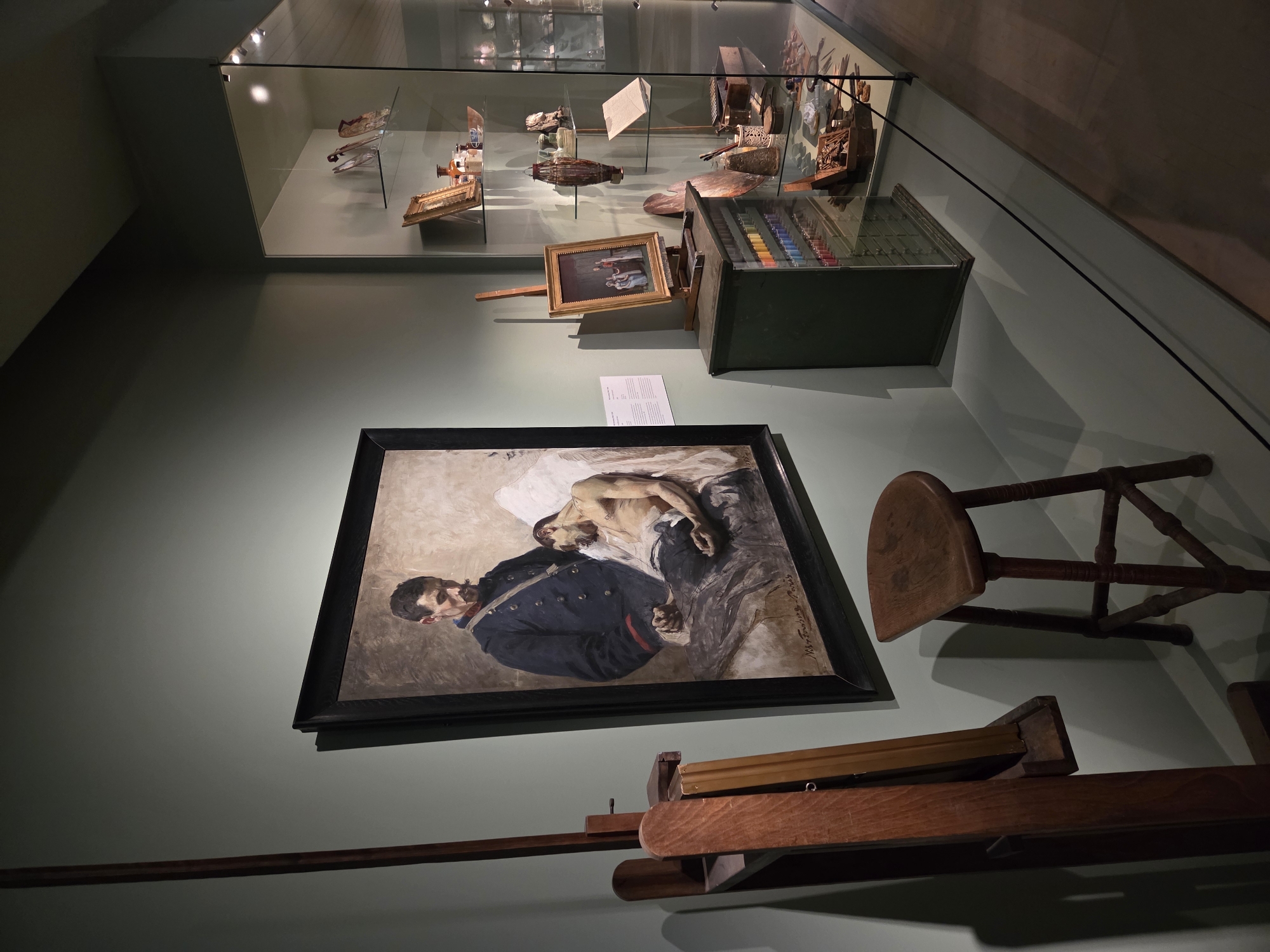 The image depicts an art exhibition, likely in Malmö, Sweden, given the context. In the foreground, a wooden easel stands to the left, next to a wooden stool. Dominating the center is a large framed painting of two men, one in military attire standing over another reclining figure. To the right, a glass display case showcases various art supplies and small decorative objects, including a cabinet filled with colorful paint tubes, brushes, palettes, and several framed artworks. The walls are painted a muted green, and the floor is a light grey. The lighting appears to be directed, highlighting the artworks and artifacts.