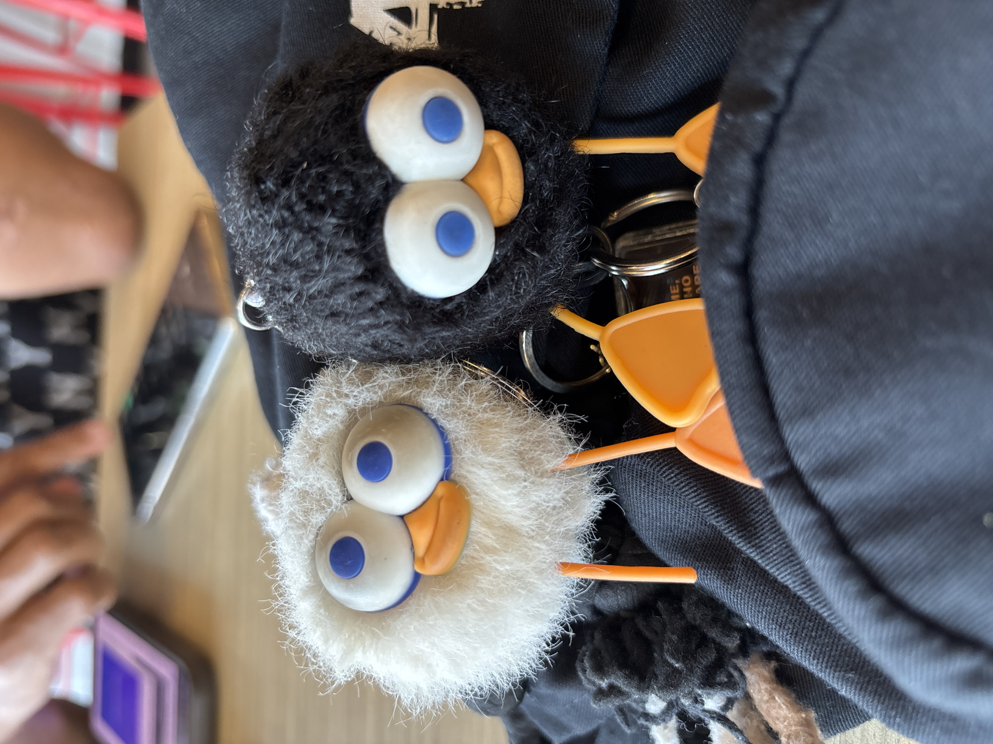 Two fluffy keychain toys, one white and one black, are placed on black fabric. Both toys have large, round, googly eyes with blue pupils and orange beaks. They also have orange stick-like legs. The white toy is on the left, and the black toy is on the right. In the background, a blurred image shows hands and objects on a wooden table, suggesting an indoor setting. The image does not provide any information about the city of Kuta Selatan, Indonesia.