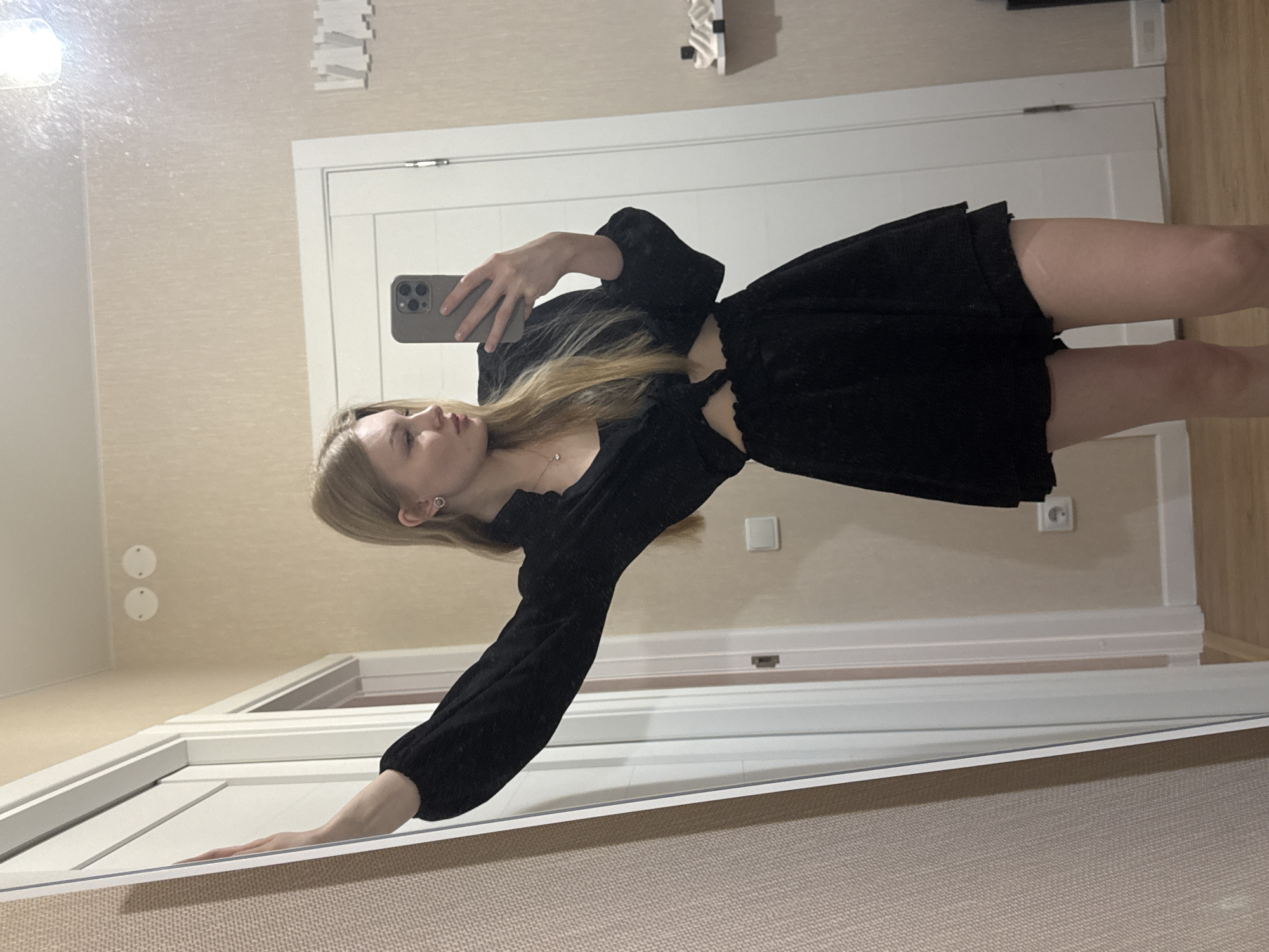 A person stands in an interior space, holding a smartphone, presumably taking a picture in front of a mirror. The individual has long blond hair and is wearing a short black dress with a long-sleeved top. The person is oriented at an angle, facing the mirror. The background features a door with white panels and trim, with parts of the interior wall visible. The setting is likely a residential interior, possibly in an urban location. There is no verifiable information to specify that the location is in Perm, Russia.
