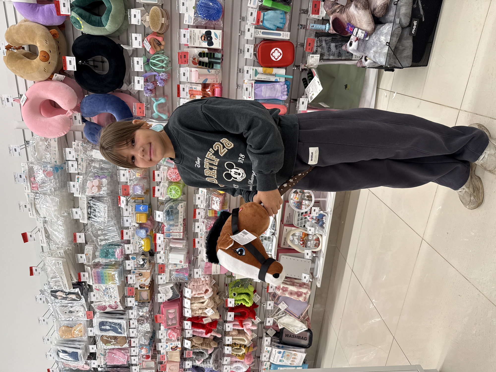 A young girl stands in a retail store, holding a stuffed horse head. She is wearing a dark gray sweatshirt with a Mickey Mouse graphic and dark gray sweatpants. The store displays a variety of products on shelves and racks, including travel pillows, cosmetic bags, and various accessories. The girl is positioned in the center of the frame, with the merchandise filling the background. The flooring appears to be tiled. The store appears to be located in Sosnovtsy, Russia.