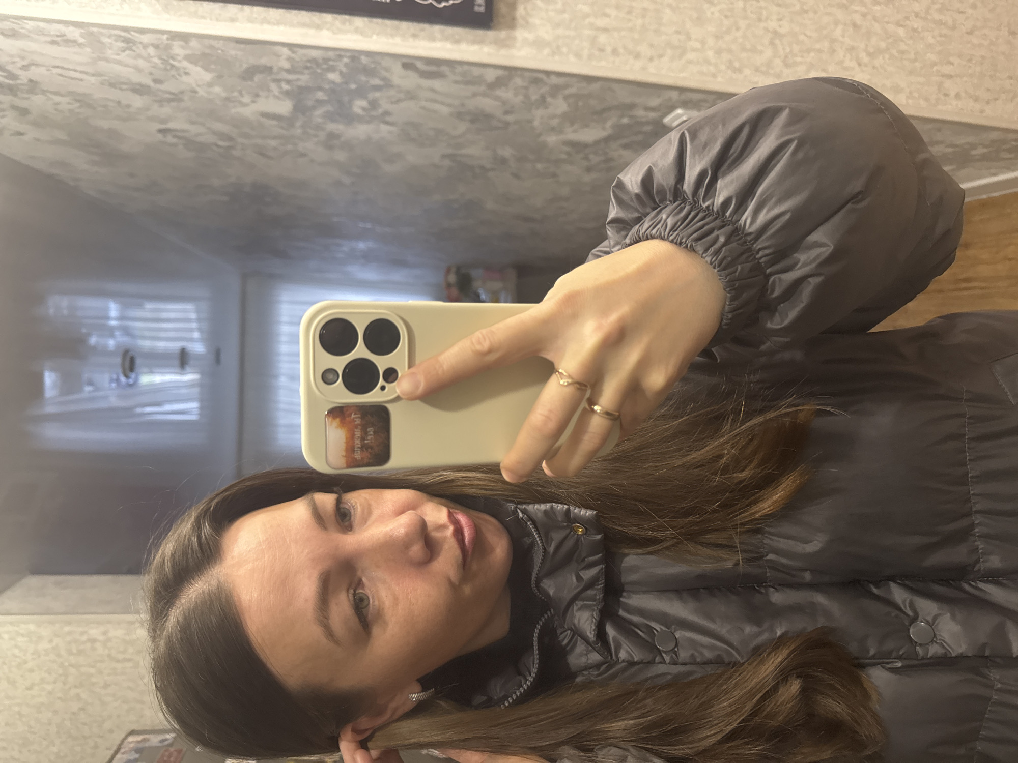 A woman in a gray puffer jacket is taking a selfie in a mirror. She holds a beige smartphone case with multiple camera lenses on its backside. Two gold rings are visible on her right hand, which is pointing toward the phone. Her hair is brown. The reflection in the mirror includes a glimpse of what appears to be an interior space with white walls and blinds. The location is not definitively identifiable as Chaikovsky, Russia, but there is no information to contradict it being there.