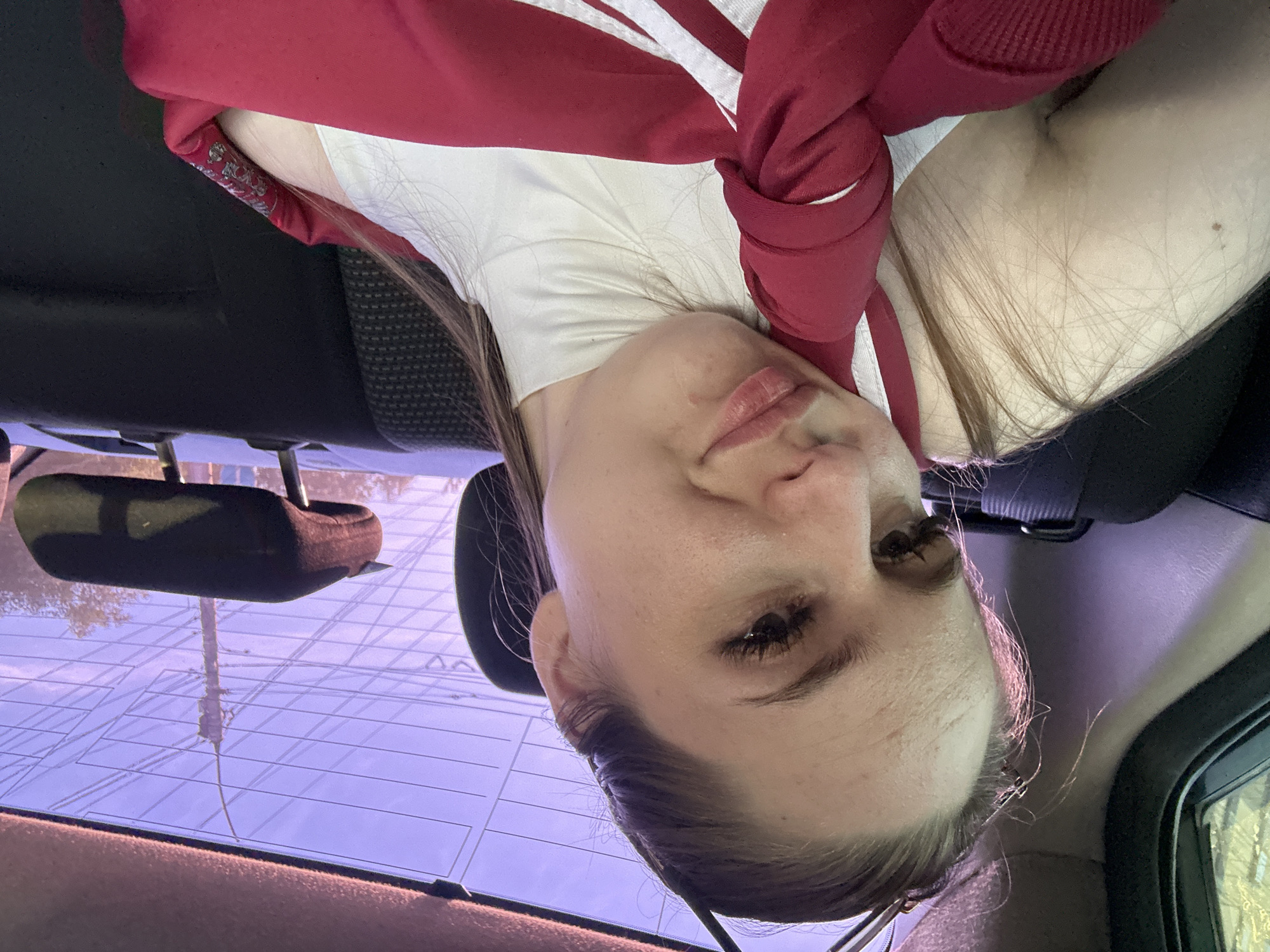 A young adult female is positioned in the back seat of a vehicle, looking directly at the camera. Her head is tilted slightly to the right, and her left hand supports her left cheek. She has light-toned skin with visible pores and minor creases on her forehead. Her brown hair is pulled back, with lighter wisps framing her face. She has prominent eyelashes. Her lips are closed, forming a slight upward curve on the left side.
She is wearing a white, high-neck top, possibly a turtleneck. Over this, a red garment with white stripes is visible, appearing to be a jacket or similar item, which is gathered or tied in the front.
The vehicle interior features dark fabric upholstery on the seats, including a headrest visible behind her and another further back on the right side of the frame. A window frame is partially visible on the left. The rear window shows a purple tint and has horizontal grid lines, likely for a defroster. Through the rear window, an exterior scene is visible: a utility pole with multiple horizontal wires, and green foliage from trees behind it. The ambient light inside the vehicle casts a pinkish-purple hue, particularly on the left side of her face and hair.