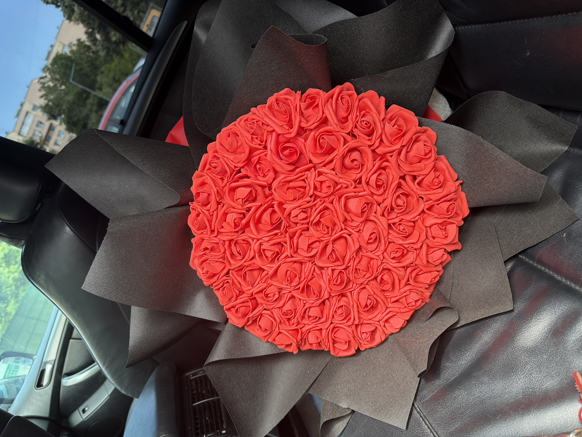A close-up, overhead shot shows a bouquet of artificial red roses, intricately arranged and wrapped in black material. The bouquet is positioned on the black leather seat of a car, likely in Pazardzhik, Bulgaria, as indicated by the external view through the car windows. Outside, a red car is visible, along with trees and buildings, suggesting an urban environment. The overall scene is focused on the bouquet, with the car's interior and the blurred exterior serving as the background.