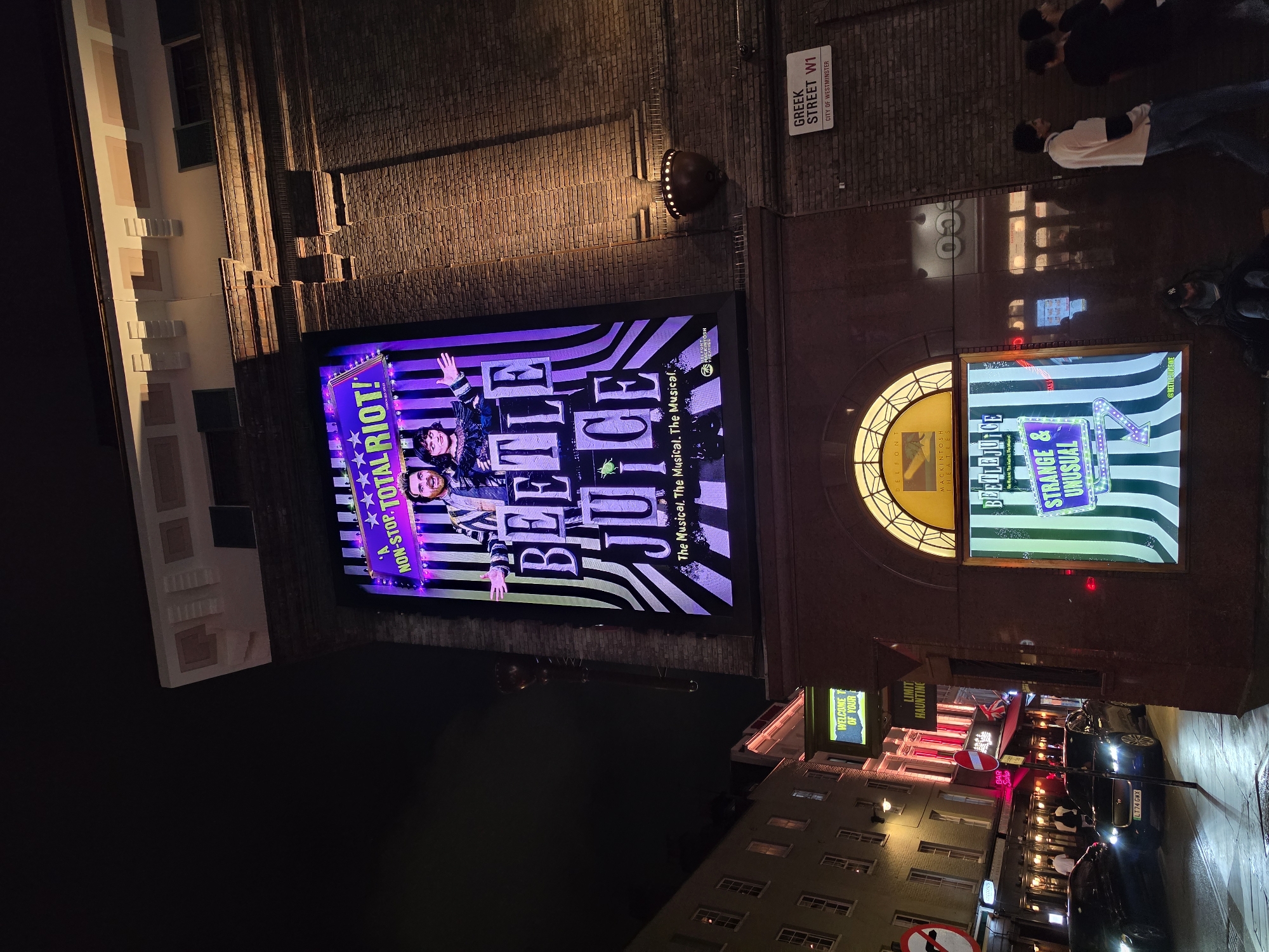 The image shows a nighttime view of a street in the Soho district of London, United Kingdom. The primary subject is a large, illuminated billboard advertising the musical "Beetlejuice" on the side of a brick building. The billboard features a stylized image of the musical's characters, the title "BEETLEJUICE," and slogans such as "NON-STOP TOTAL RIOT!" and "STRANGE & UNUSUAL." Below the main billboard, a smaller illuminated display also advertises the musical.
In the foreground, a dark-colored car is parked on the street, and a street sign indicates "GREEK STREET W1 CITY OF WESTMINSTER." Further down the street, other buildings with illuminated windows and signs are visible, suggesting a bustling commercial area. Several pedestrians are seen walking on the sidewalk. The overall scene is characteristic of a vibrant entertainment district at night.