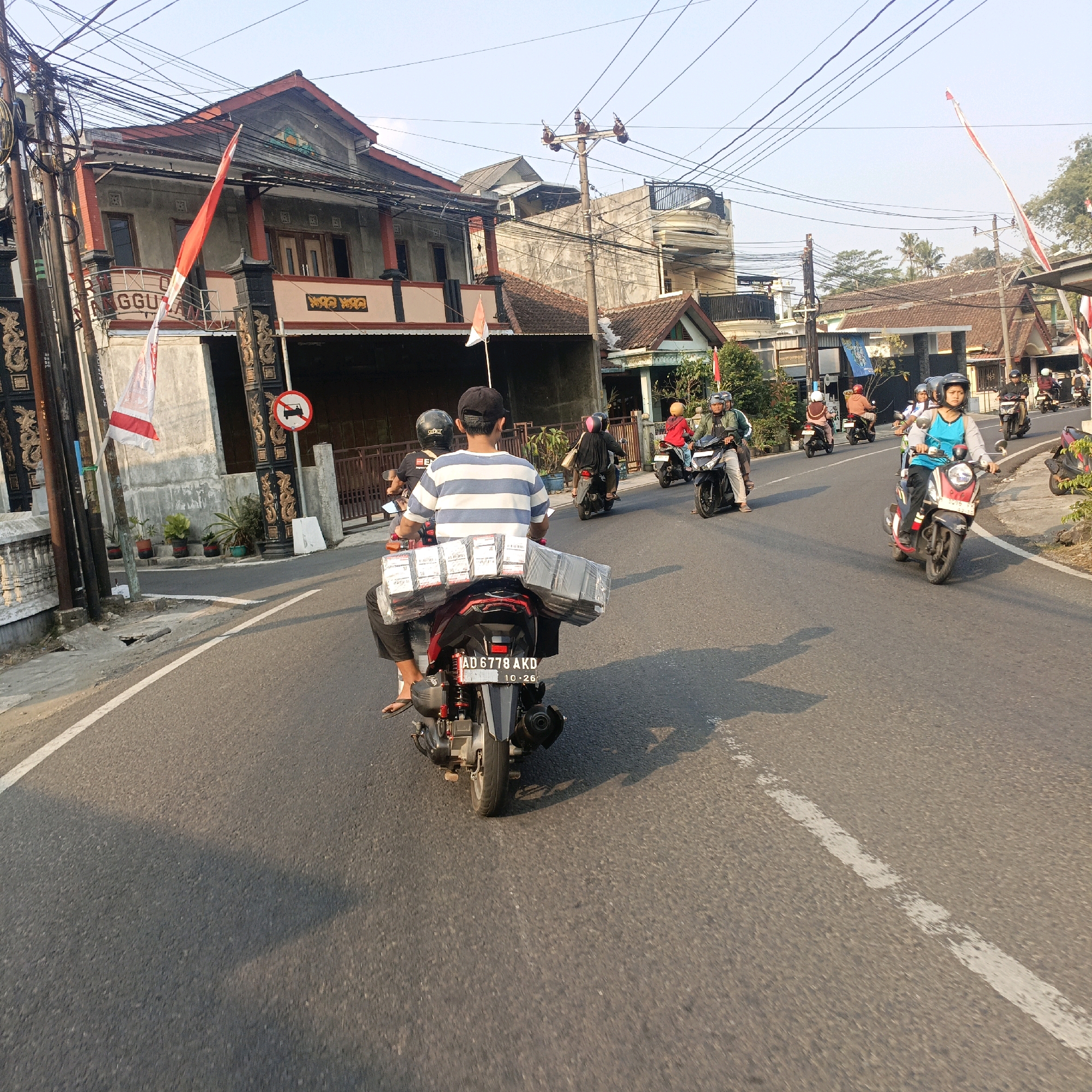 In Salatiga, Indonesia, on a paved road bathed in the warm glow of late afternoon sun, a procession of motorbikes navigates through a residential area. The air is filled with the distant hum of engines and the rustle of Indonesian flags, their red and white colors fluttering from poles and draped across buildings.
In the foreground, a motorbike carries a driver and a passenger, the latter a man in a striped t-shirt, his back to the camera. Strapped to the back of their motorbike are numerous rectangular boxes, neatly stacked and wrapped in clear plastic, suggesting a delivery. The motorbike's license plate reads "AD 6778 AKD".
Further ahead, a woman on a motorbike, wearing a blue top and a black helmet, leads a line of riders. Behind her, several other motorbikes follow, their riders also wearing helmets, indicating adherence to safety regulations. Buildings line the street, their architecture varied, from traditional Indonesian styles with tiled roofs to more modern structures. Utility poles, adorned with a complex web of wires, stand tall against the clear blue sky, a common sight in Indonesian urban landscapes. Lush green trees and potted plants add a touch of nature to the scene, softening the hard edges of the urban environment. The overall atmosphere is one of everyday life unfolding on a typical Indonesian street, with a sense of movement and activity.
