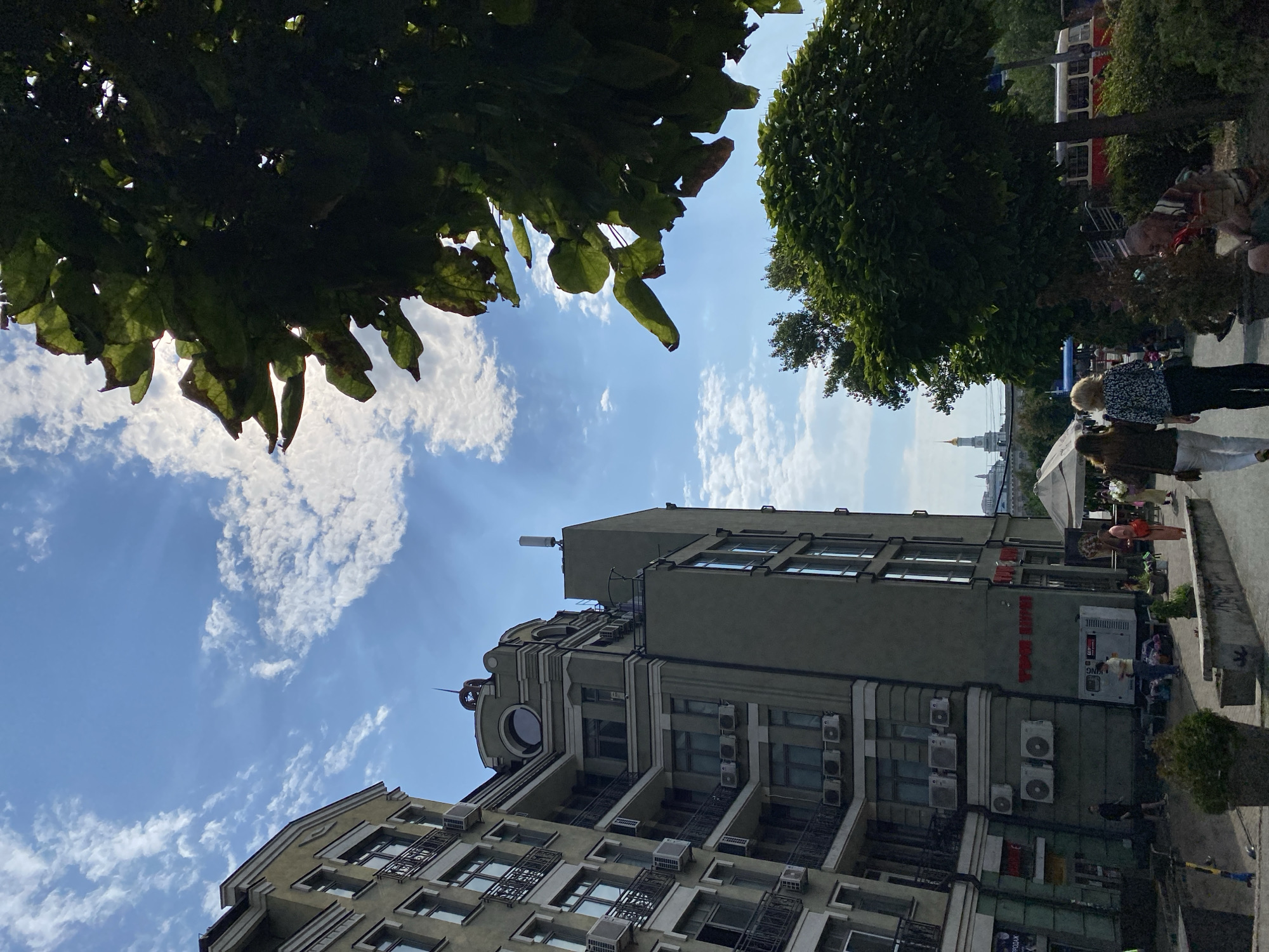 The image depicts an outdoor urban scene with several multi-story buildings. The sky is blue with scattered white clouds. In the foreground, to the right, is a large tree with dark green leaves, partially obscuring the view. A smaller, ornamental tree is also visible. To the left are two large, modern apartment buildings with balconies and numerous air conditioning units. A street or walkway runs between the buildings and the trees, with several people walking and standing. Some of the people are carrying bags. In the middle ground, a white tent is set up, and a large generator is visible near one of the buildings. A red and white tram is parked further down the street. In the background, a distant building with a golden dome and spire can be seen. The overall impression is of a sunny day in a city with a mix of residential and commercial architecture, with some street-level activity.