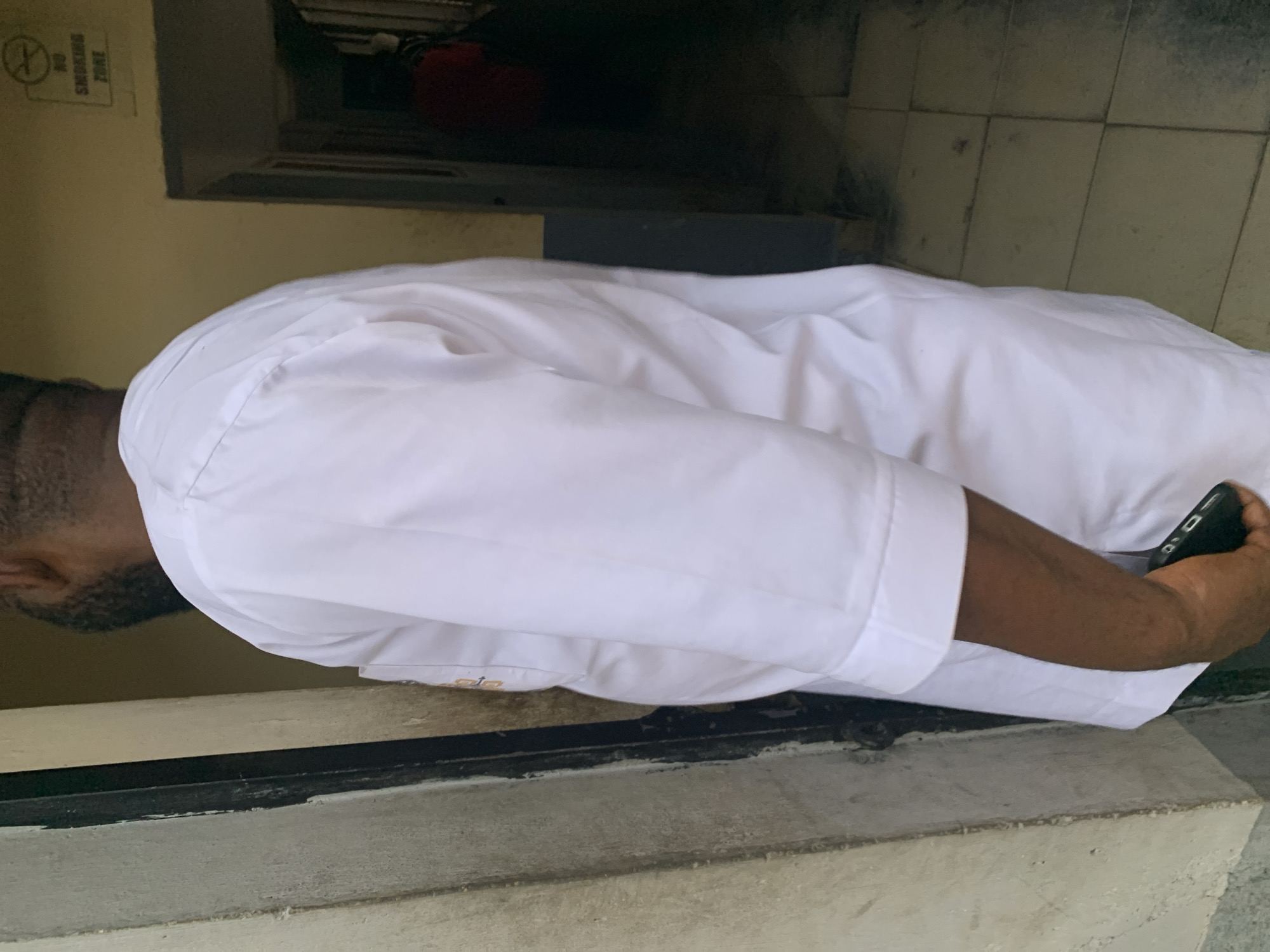 A man wearing a white shirt is looking downwards. The background shows a "No Smoking Zone" sign and a tiled wall. The location is New Stadium, Nigeria. The man is holding a phone in his right hand.