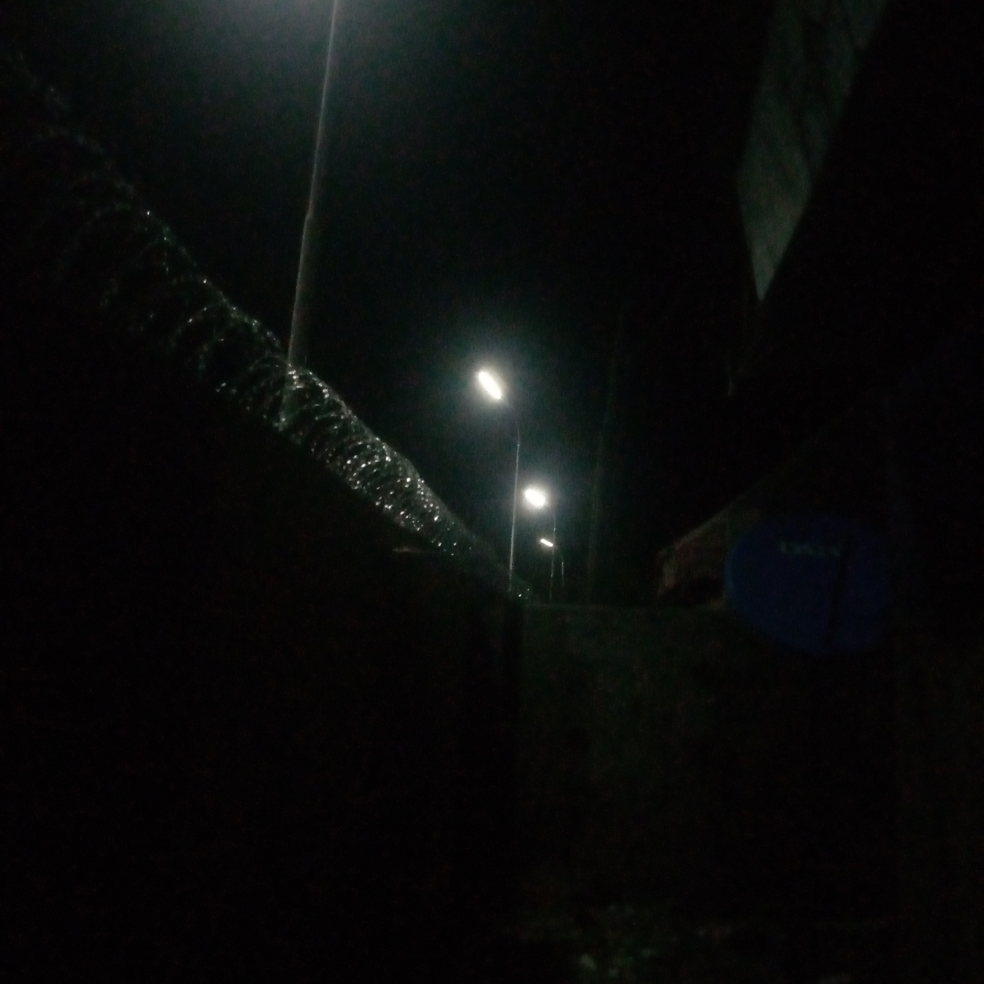 An image captured at night shows a chain-link fence topped with barbed wire, illuminated by streetlights. The fence runs diagonally across the frame, from the upper left to the lower right. Behind the fence, several streetlights are visible, casting a glow on the dark scene. One streetlight is positioned in the mid-ground, and two more are further back. A dark, rectangular object, possibly a satellite dish, is visible on the right side of the frame. The overall environment is dark and appears to be an outdoor area in a city.