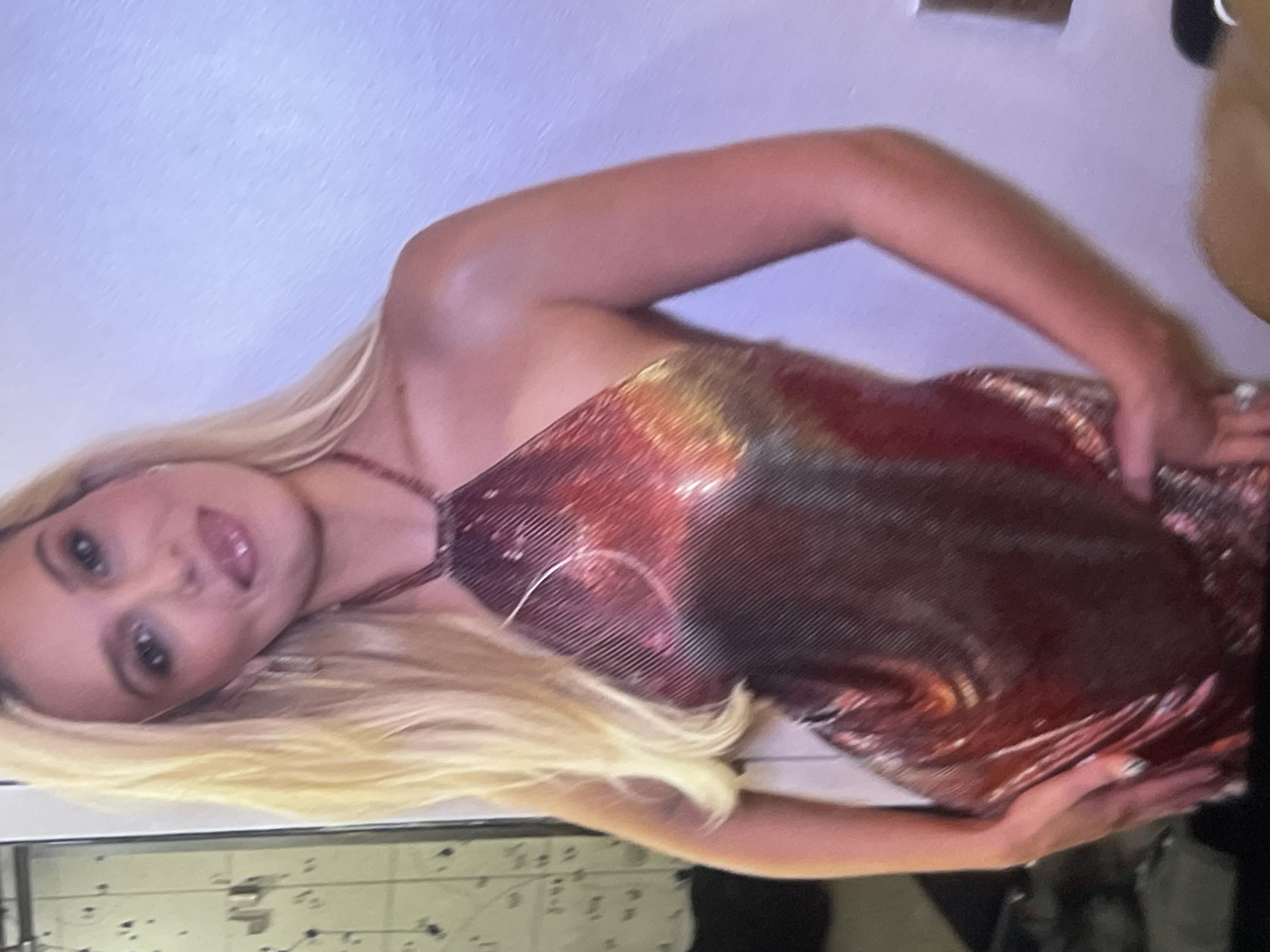 A young woman with long blonde hair and fair skin is captured in a portrait-style photograph, likely taken indoors in Poltava, Ukraine. She is depicted from the waist up, with her head slightly tilted, looking directly at the camera with a subtle, pleasant smile.
She is wearing a striking, iridescent halter-neck top or dress that shimmers with colors transitioning from golden and coppery hues at the top to deeper reds and maroon towards the bottom. The fabric appears to be a mesh or sequined material, creating a textured, reflective surface. A thin, dark strap, possibly part of the garment, is visible around her neck. Her arms are bent, with her hands resting on her hips or the sides of her dress. She wears rings on her fingers, and her fingernails are painted.
The background is a plain, light-colored wall, possibly white or a very light pastel. To the bottom left, a dark, horizontal surface with some indistinct markings or objects is visible. The lighting is artificial, enhancing the reflective qualities of her attire and highlighting her skin. The image suggests a posed moment, possibly at an evening event or social gathering. No visible text is present in the image.