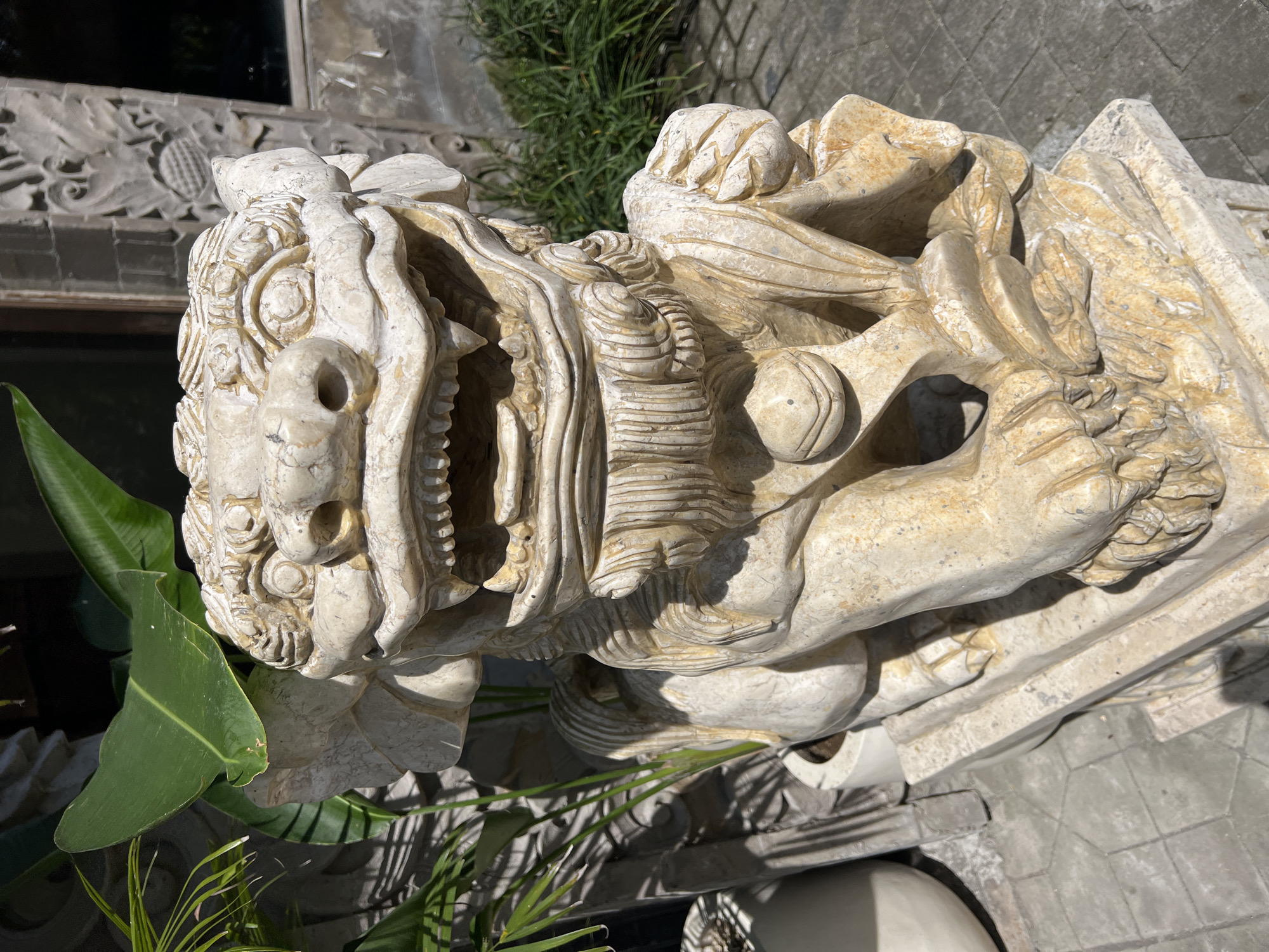 A pale stone statue of a mythical beast with an open mouth and sharp teeth stands outdoors. The sculpture is intricately detailed, with textured fur-like patterns. The beast's nose has prominent holes. Foliage flanks the statue on the left. The background includes a building with carved decorative elements and surrounding greenery. The statue rests on a stone base. The setting appears to be the city of Petulu, Indonesia.