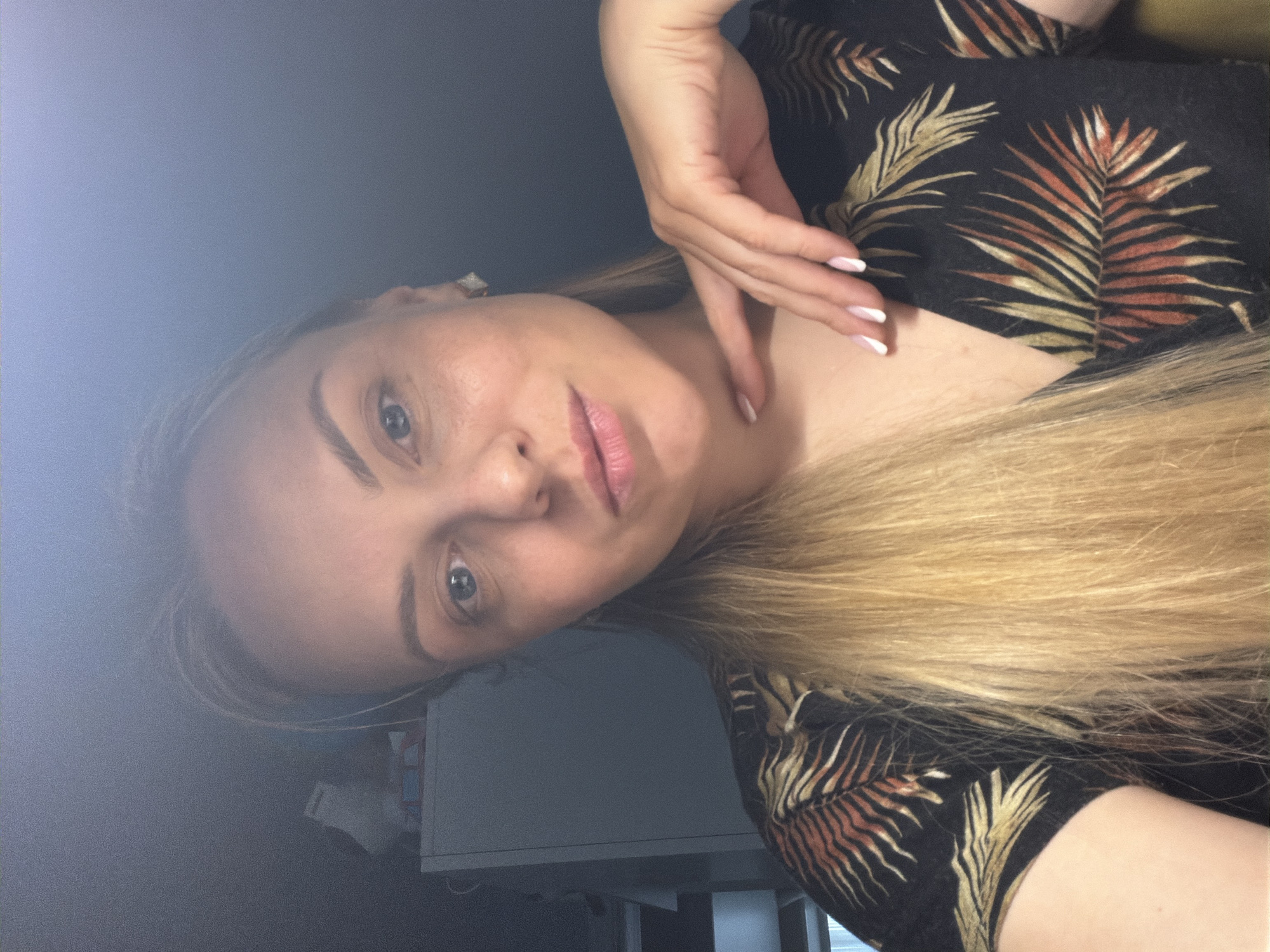 A light-skinned woman with blonde hair is posing for a selfie indoors. She is wearing a black top with a fern print in shades of brown and gold. Her left hand is raised and touching her neck. Her fingernails are painted with white tips. The background is blurred, but a white cabinet is partially visible. The photograph appears to be taken in a room with dim lighting. There is no indication of the city of Votkinsk, Russia.